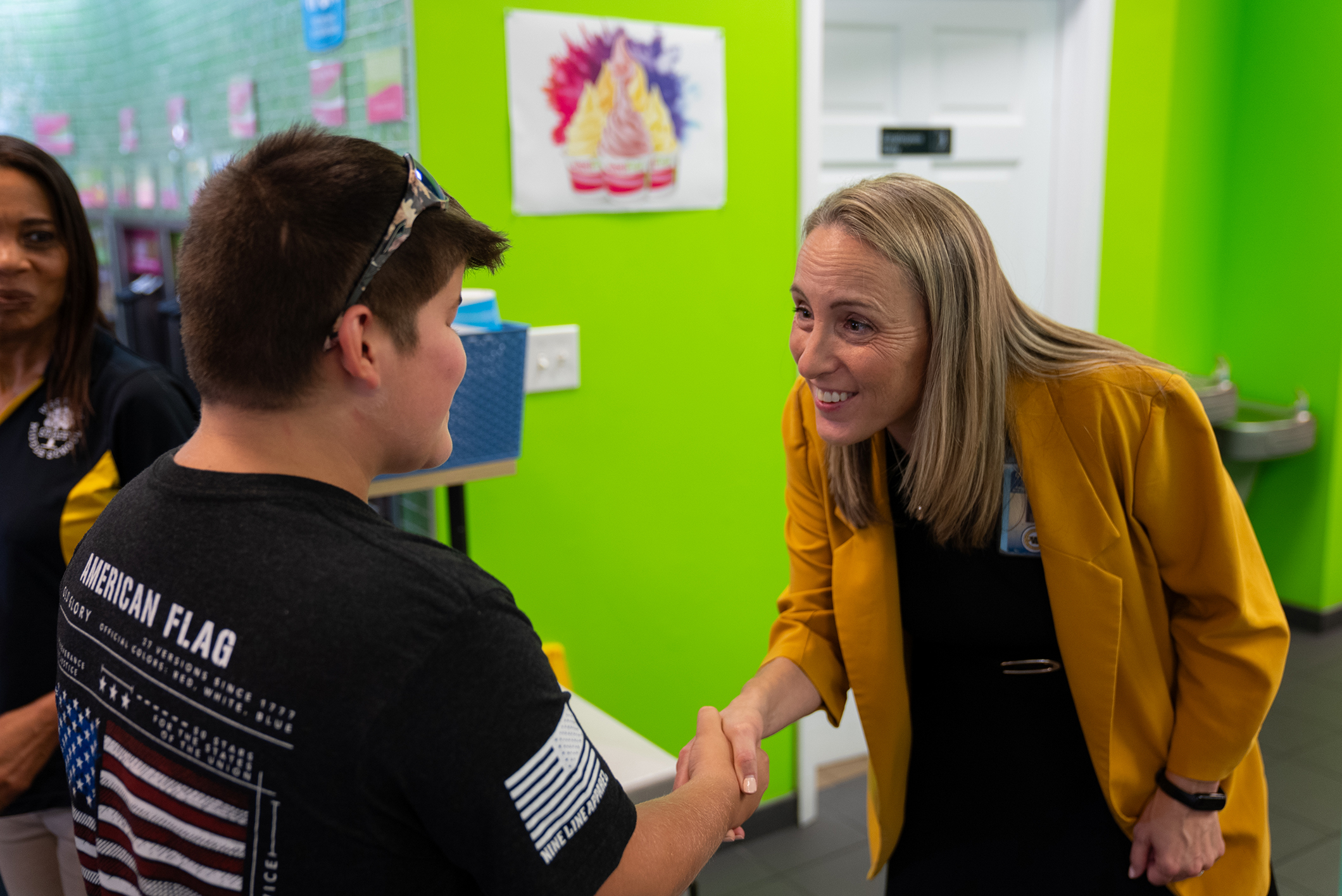 A man in a black shirt shakes hands with a woman in a yellow jacket. Behind them is a green wall with a white door.