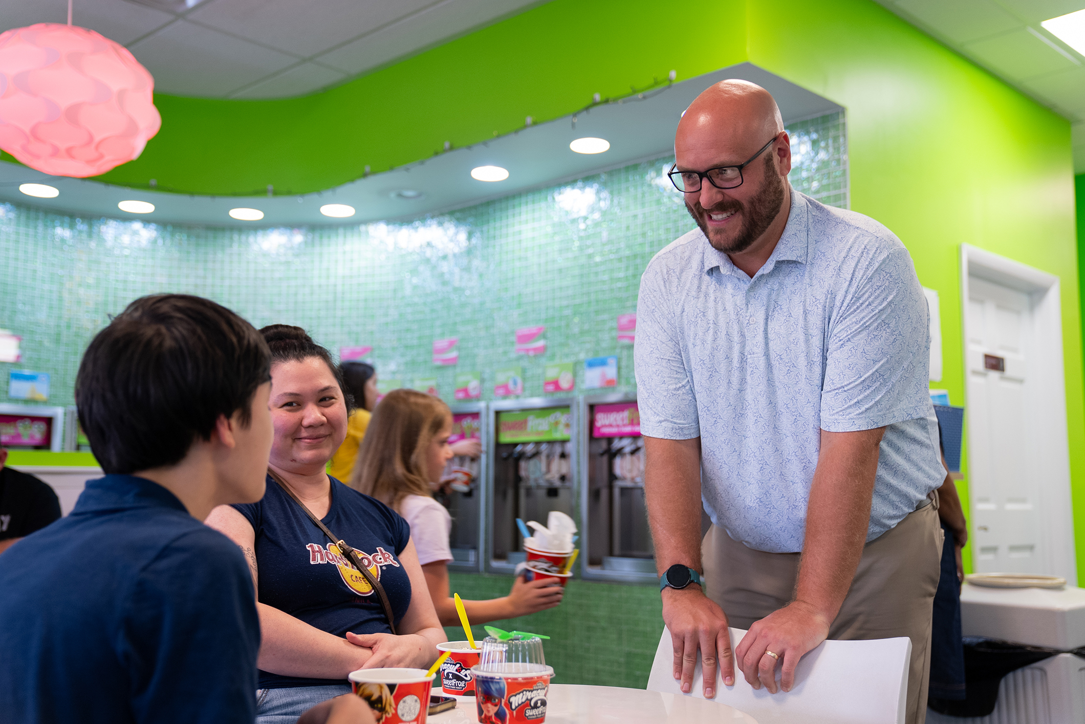 A man stands at a table in a brightly lit eatery, smiling, with a group of people seated.