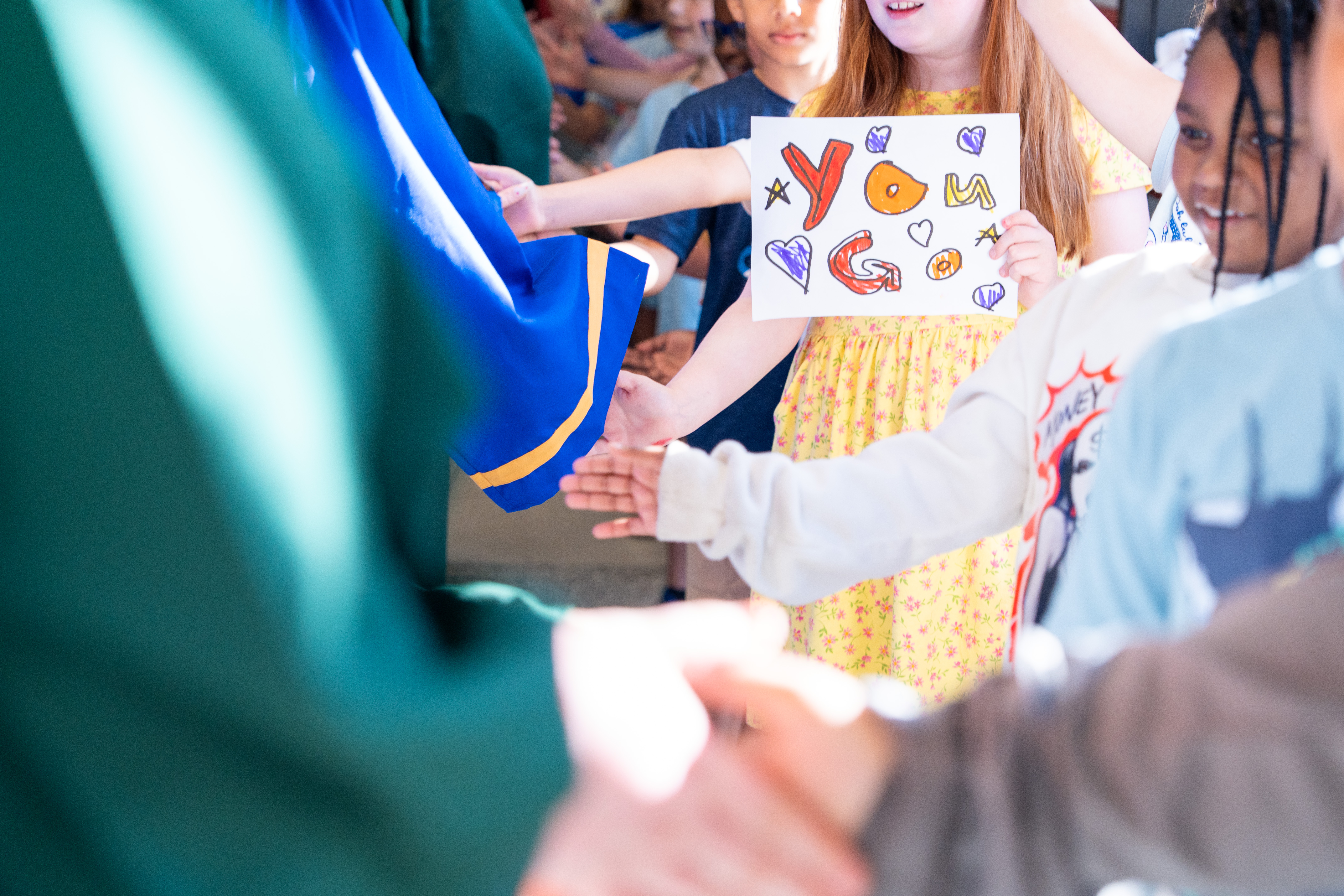 Several kids hold hands, a girl holds a colorful sign. They seem to be gathered in a classroom.