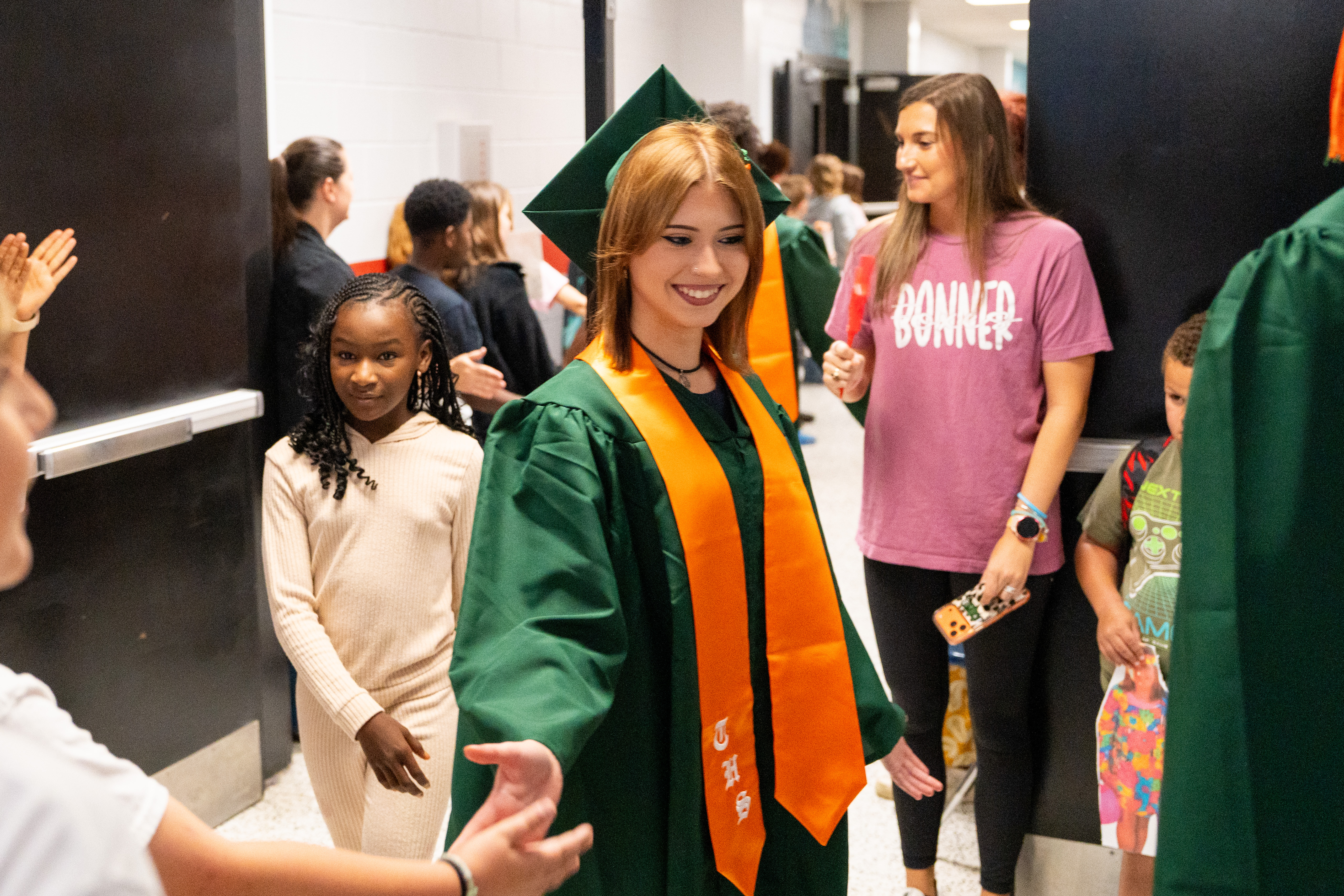 A person in a green gown walks with an orange sash, smiling. Nearby, others clap.