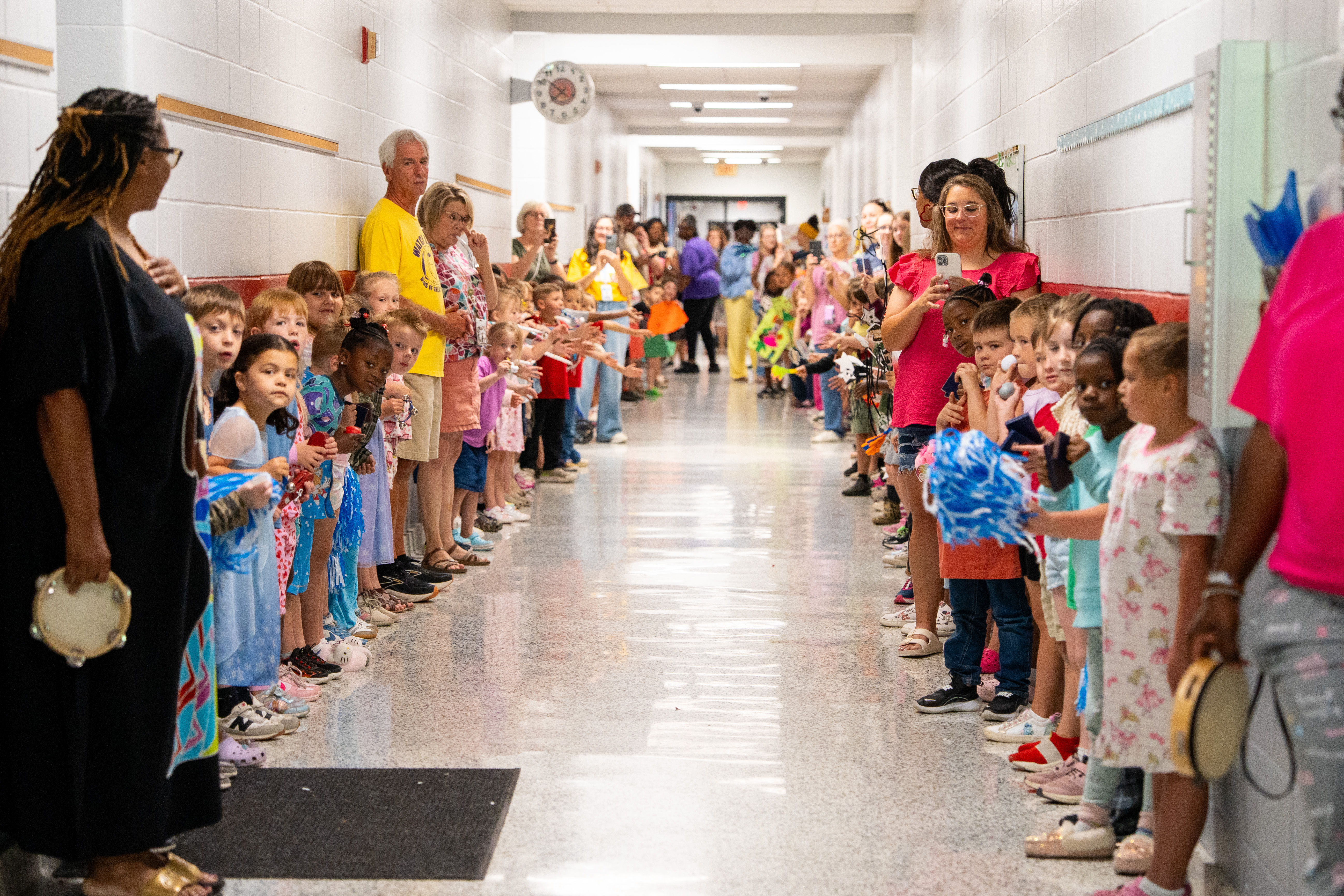 School corridor with children lined up, some holding objects, accompanied by adults; walls have art and a clock.