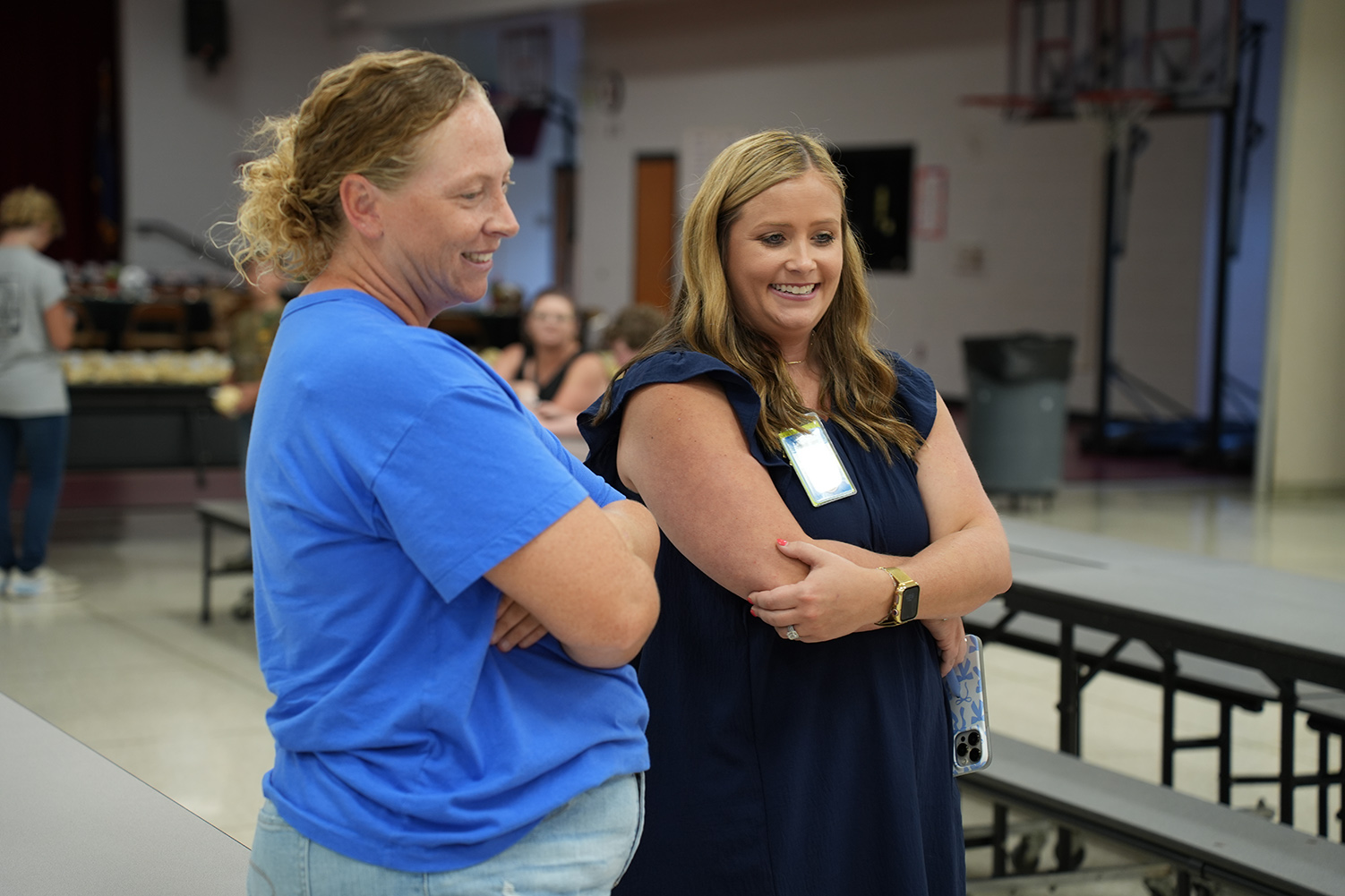 Two women converse in a room with tables, benches, and a basketball court in the background.