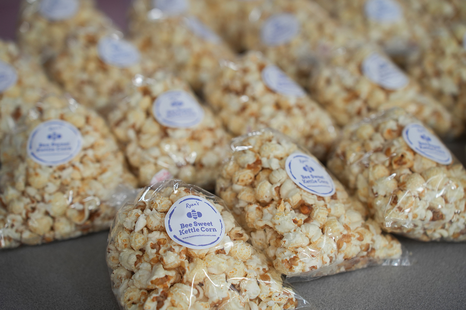 Numerous bags of popcorn with white labels displaying text and a logo sit on a gray surface.