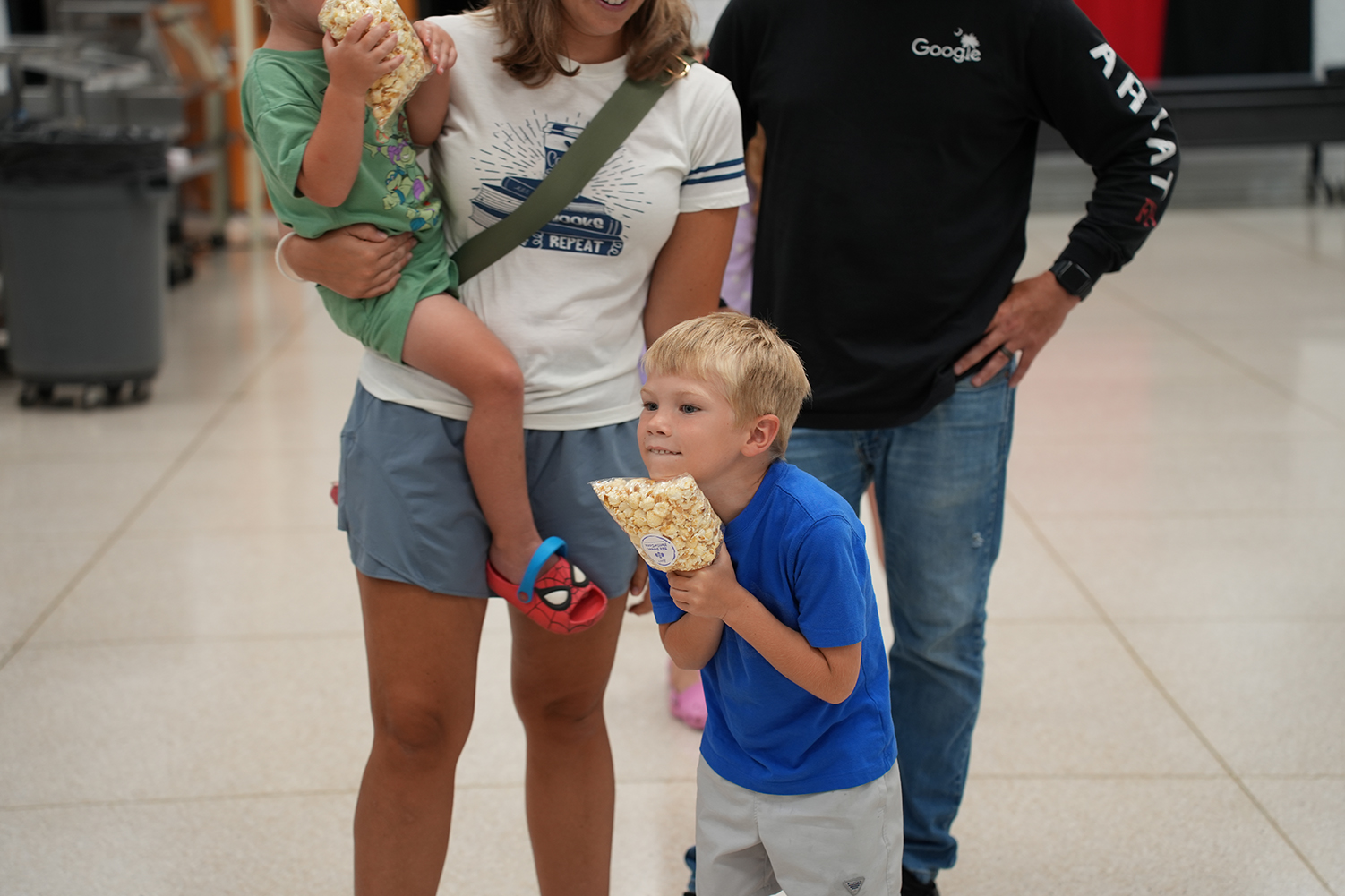 A woman holds a child in one arm while a boy eats popcorn. Another boy stands beside them, also eating popcorn. A man is behind them, holding his hand on his hip.