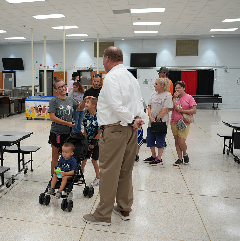 A group of adults and children in a hall with white tile floor. A man with a watch speaks to a young boy in a stroller.