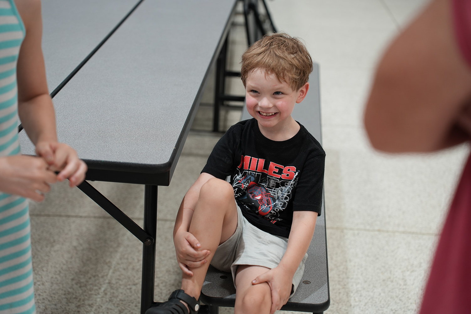 A boy with short hair and a smile sits on a bench, wearing a black t-shirt.