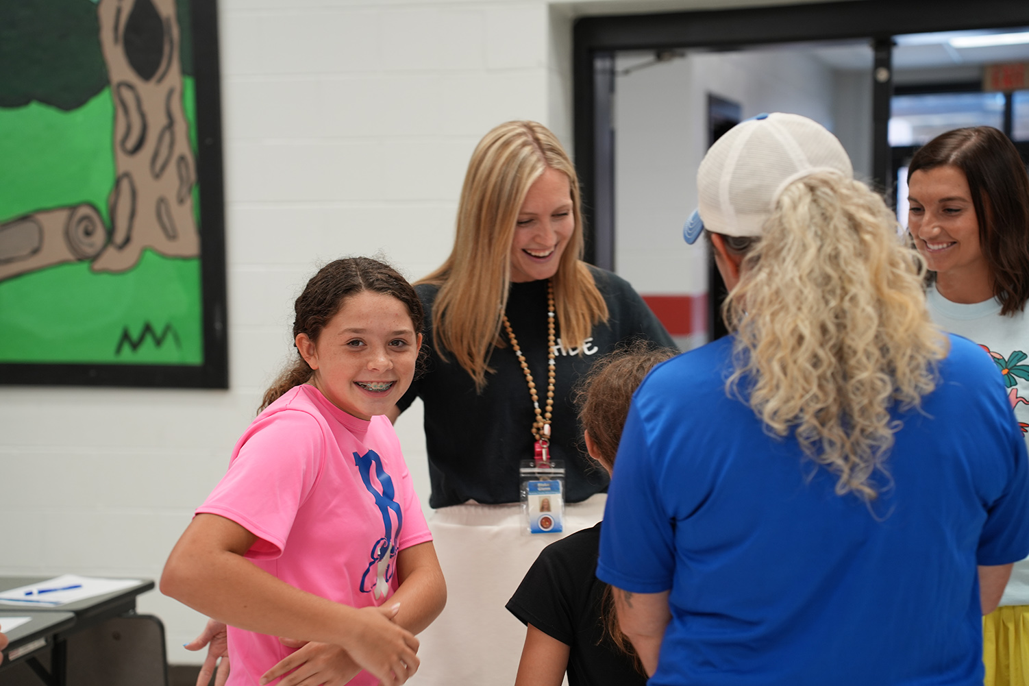 Several people stand in a room; a young girl with a pink shirt stands with a woman in black, and a man with a blue hat.