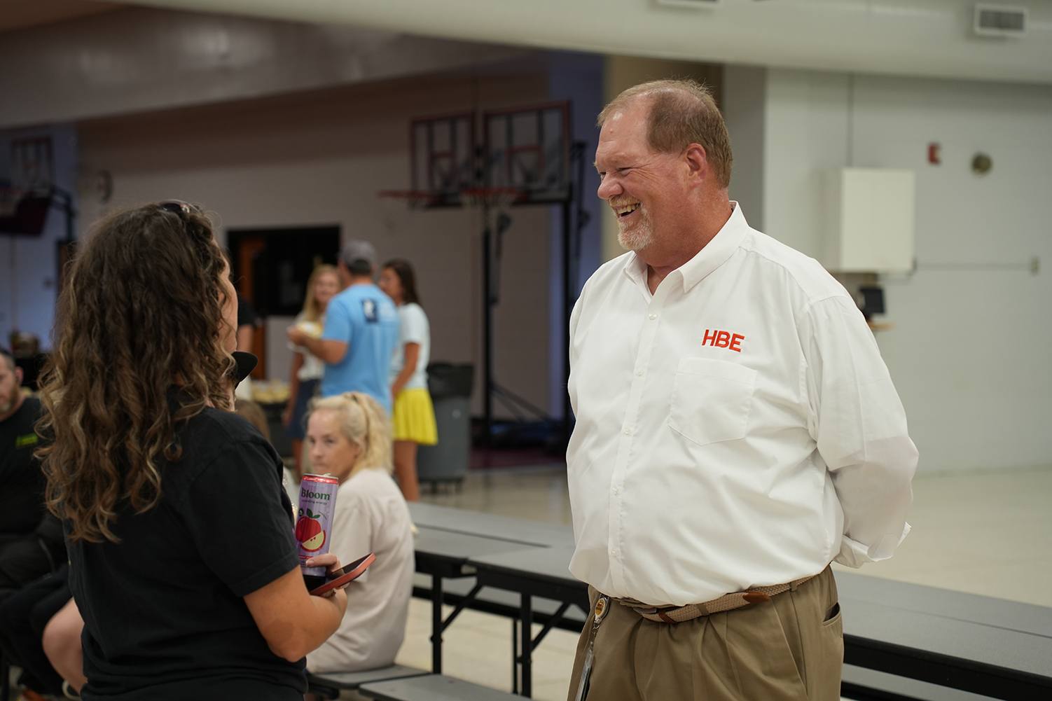 A man in a white shirt with a logo talks to a woman in a black shirt. They are in a room with tables and a basketball court in the background.