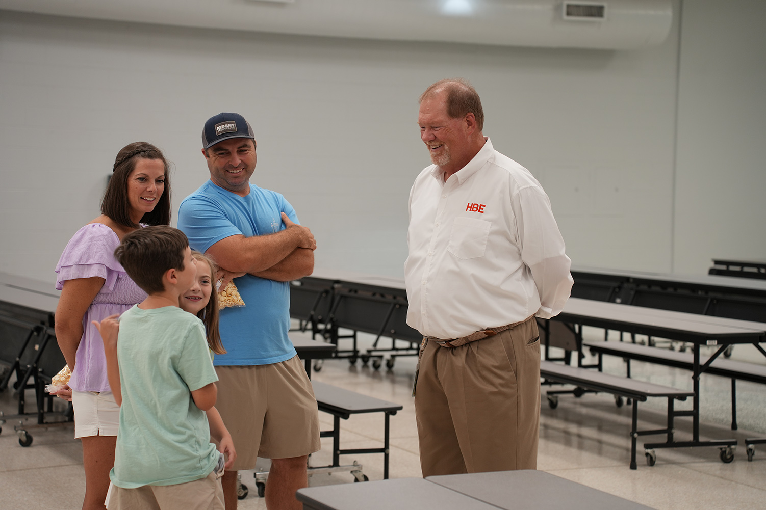 Four people stand in a room with folding tables and chairs. Two adults are smiling and speaking.