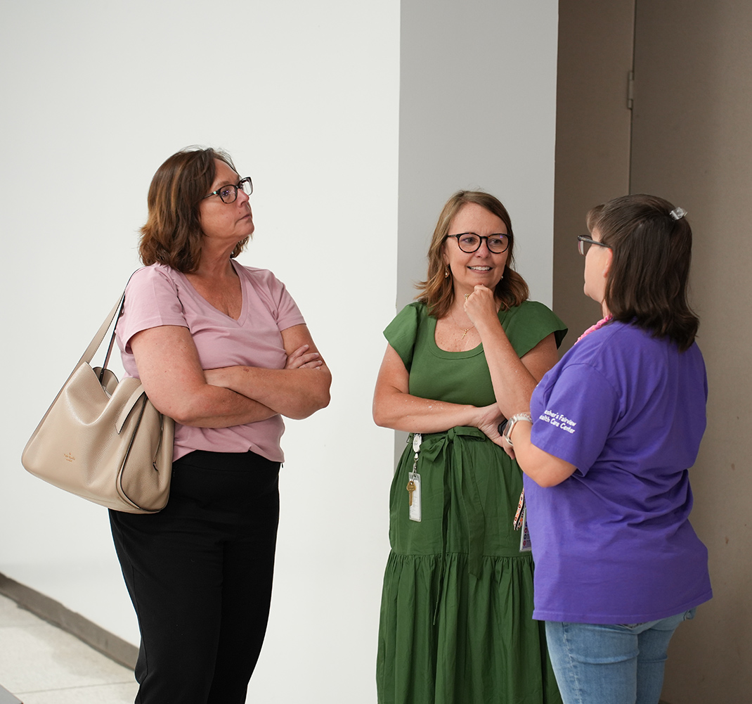 Three women in an office corridor. One wears a pink shirt and black pants, another green with a belt, and the third purple with glasses.