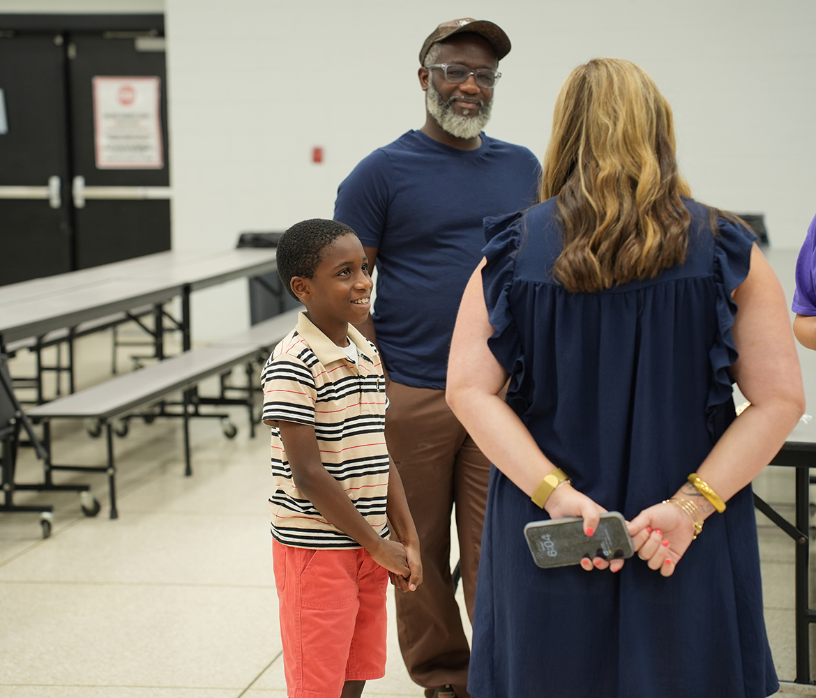 A family with a young child stands in a room with white walls. The child looks at the camera.