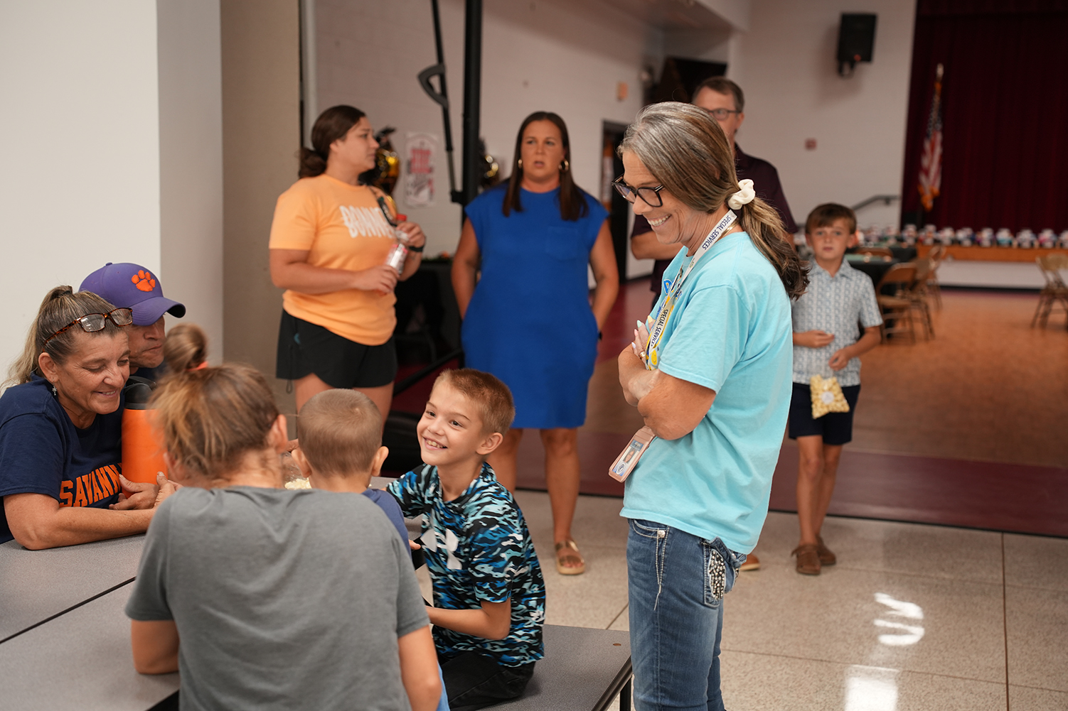 A group of people in a room. A woman in a blue shirt stands with a child. Others sit or stand.
