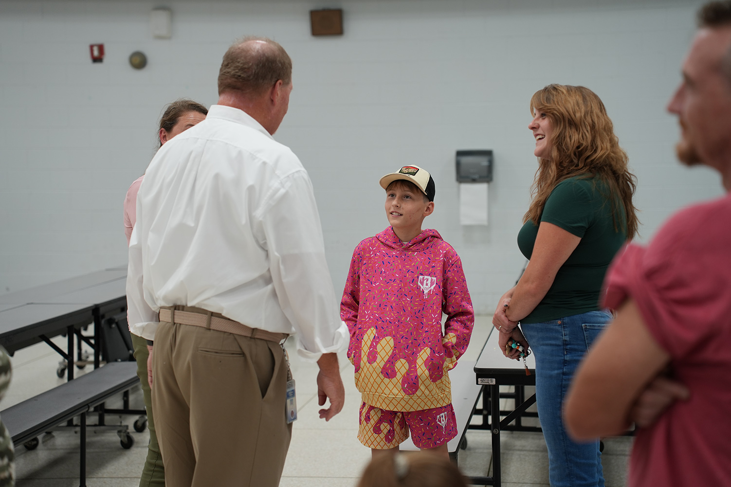 A group of people stand in a room with white walls, including a boy in pink and yellow shorts.