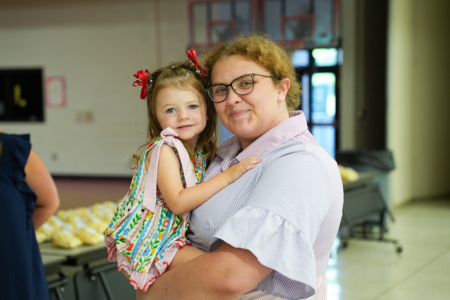 Woman wearing glasses and a pink shirt holds a young girl with red bows in her hair.