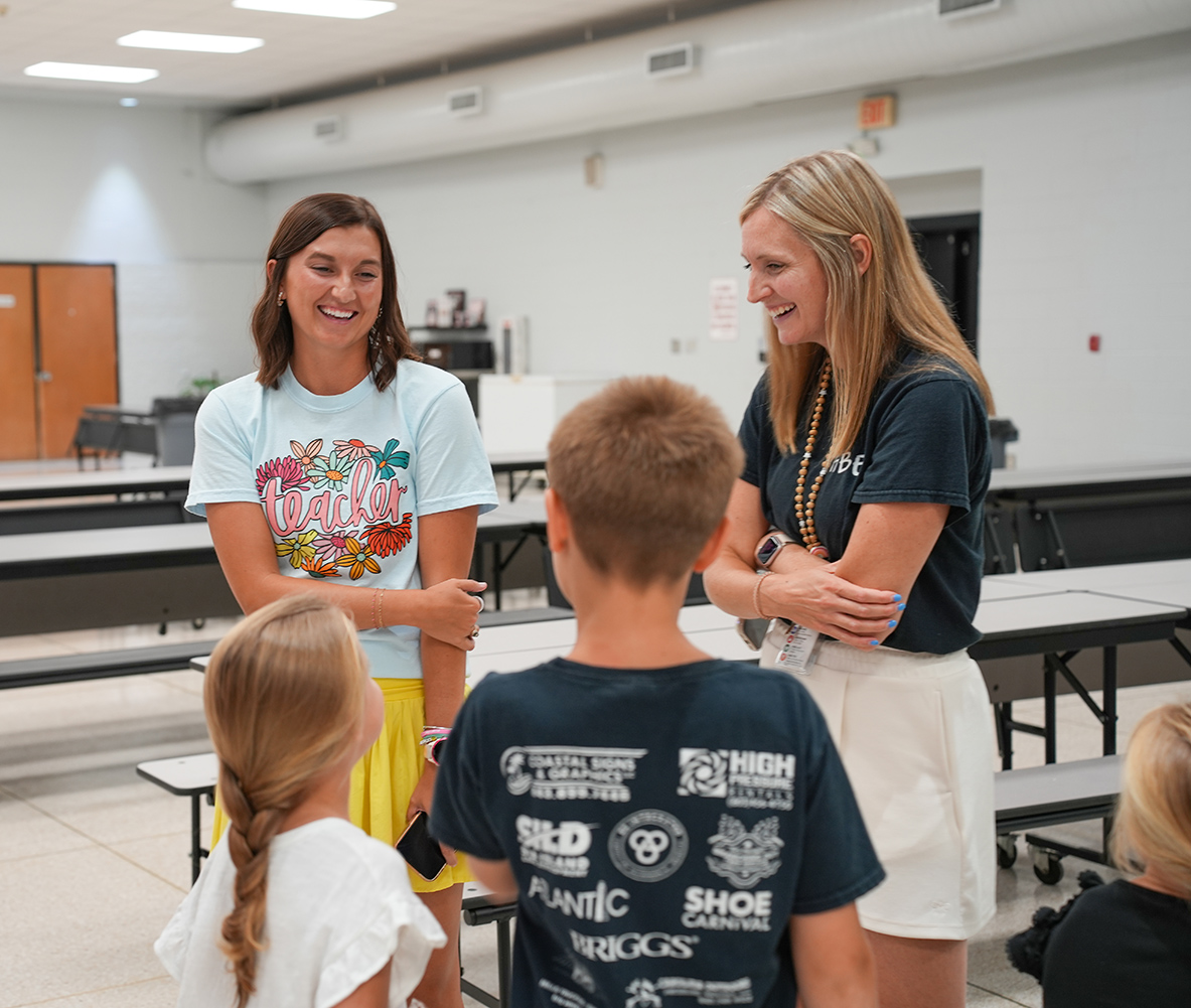 Two women and two children stand in a classroom. One woman smiles at the boy. The other woman wears a necklace and has arms crossed.