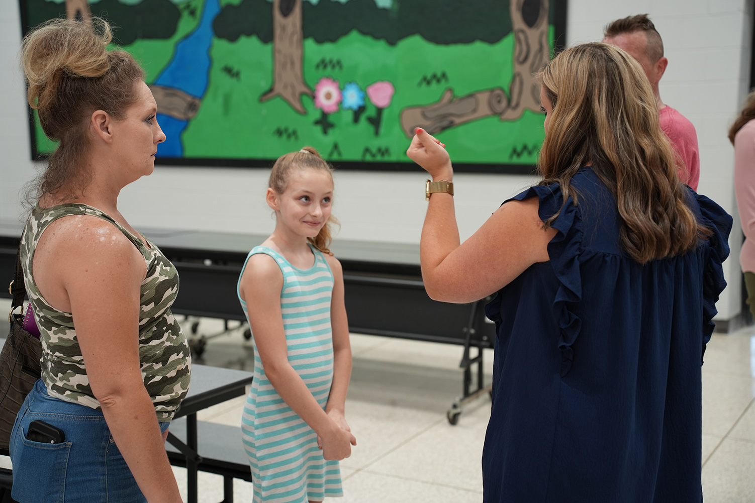 Two women and a young girl stand in a room. One woman holds a sign. Behind them, a colorful mural is on the wall.