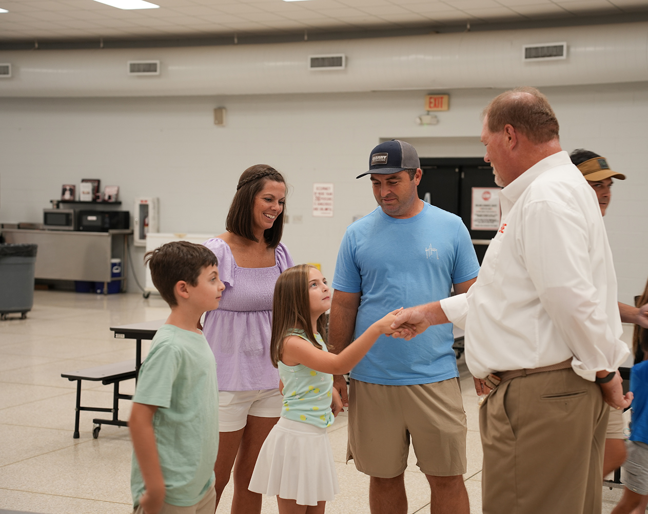 A man and woman shake hands with a boy and girl inside a white room with fluorescent lights.