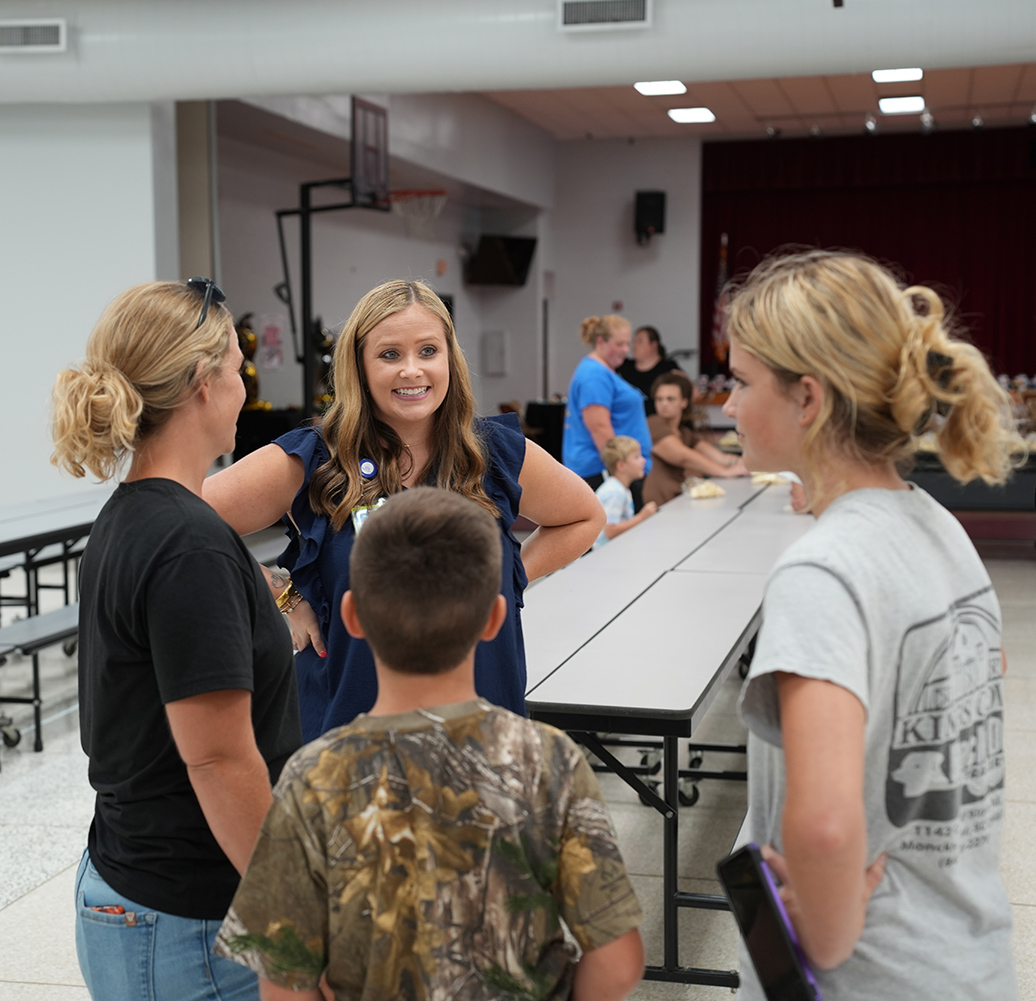 Four people conversing in a cafeteria; a woman in a blue dress, a boy in camouflage, and two women.