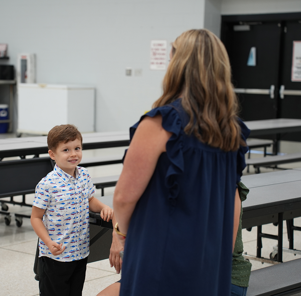 A woman with blonde hair in a blue dress talks to a boy in a white shirt. They are in a room with tables and chairs.