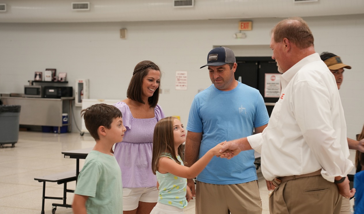 A group of people in a room. Two adults shake hands with a child. A girl in a white skirt holds the man's hand.