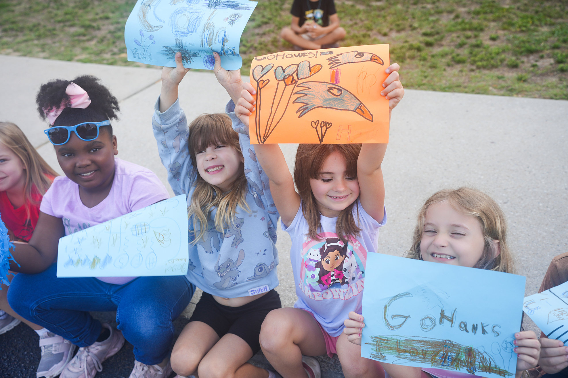 Children sit in a circle, holding paper signs. One holds a drawing of a bird. A grassy area is in the background.
