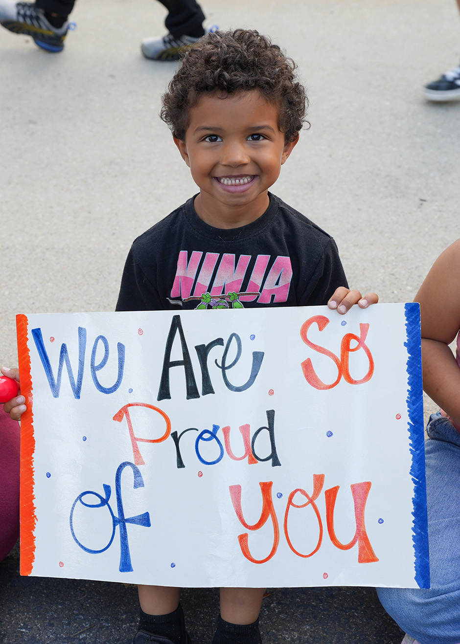 A child holds a sign that says "We Are So Proud of You" with blue and orange text on a white background.