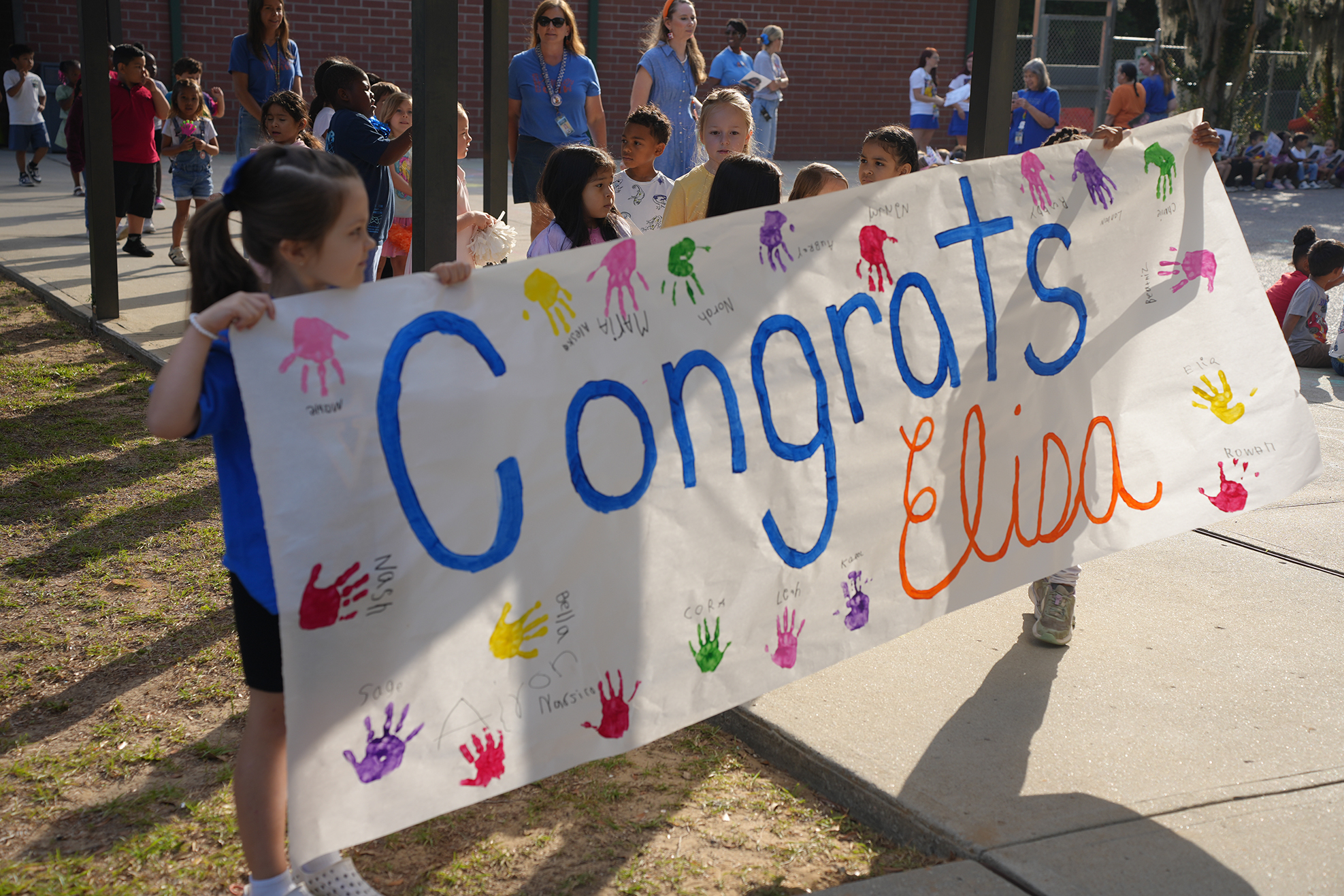 Children hold a banner with painted hands and "Congrats" and "Elina" written in blue and orange.