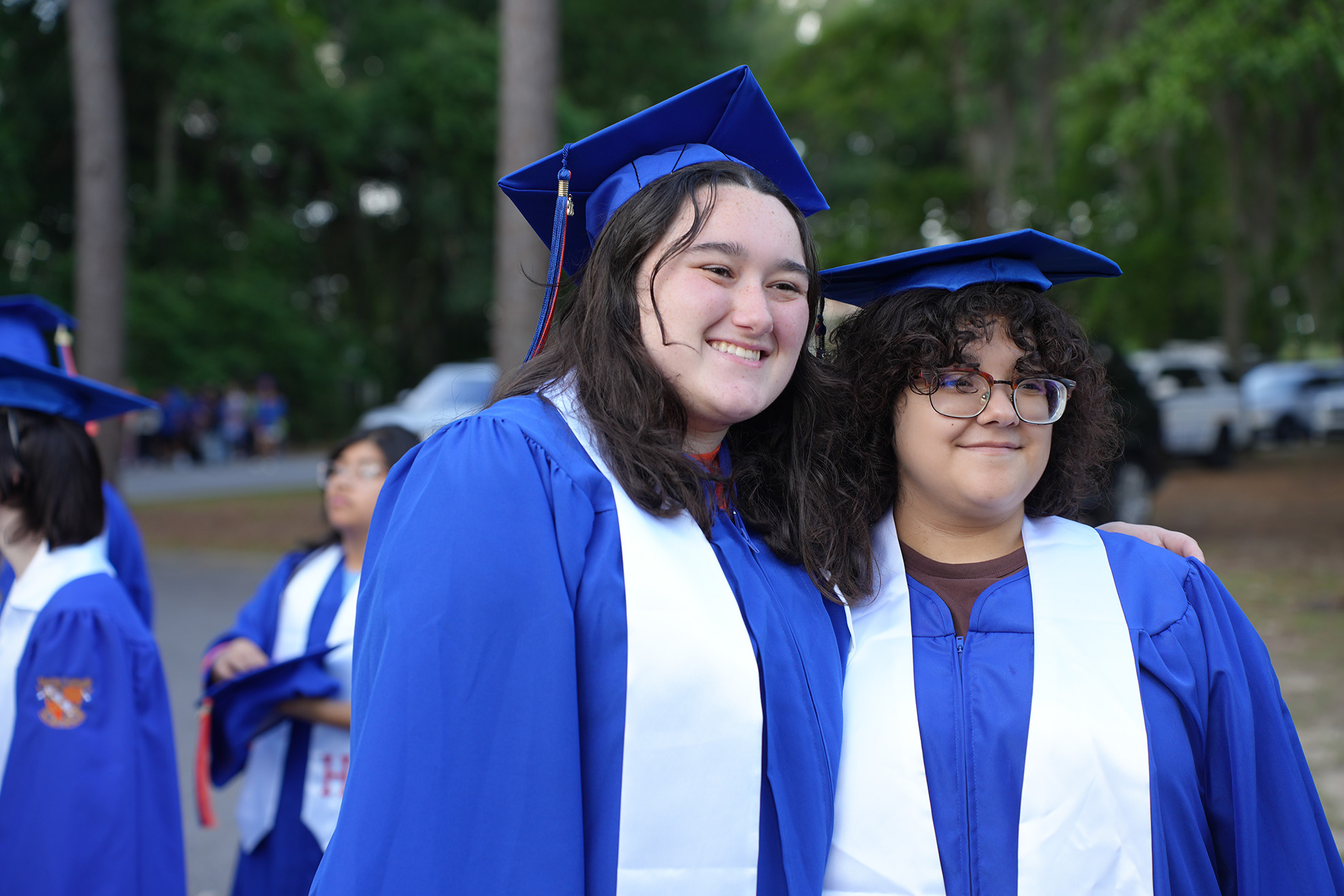 Two women in blue graduation gowns and hats smile for a photo. They stand near other students, with greenery in the background.