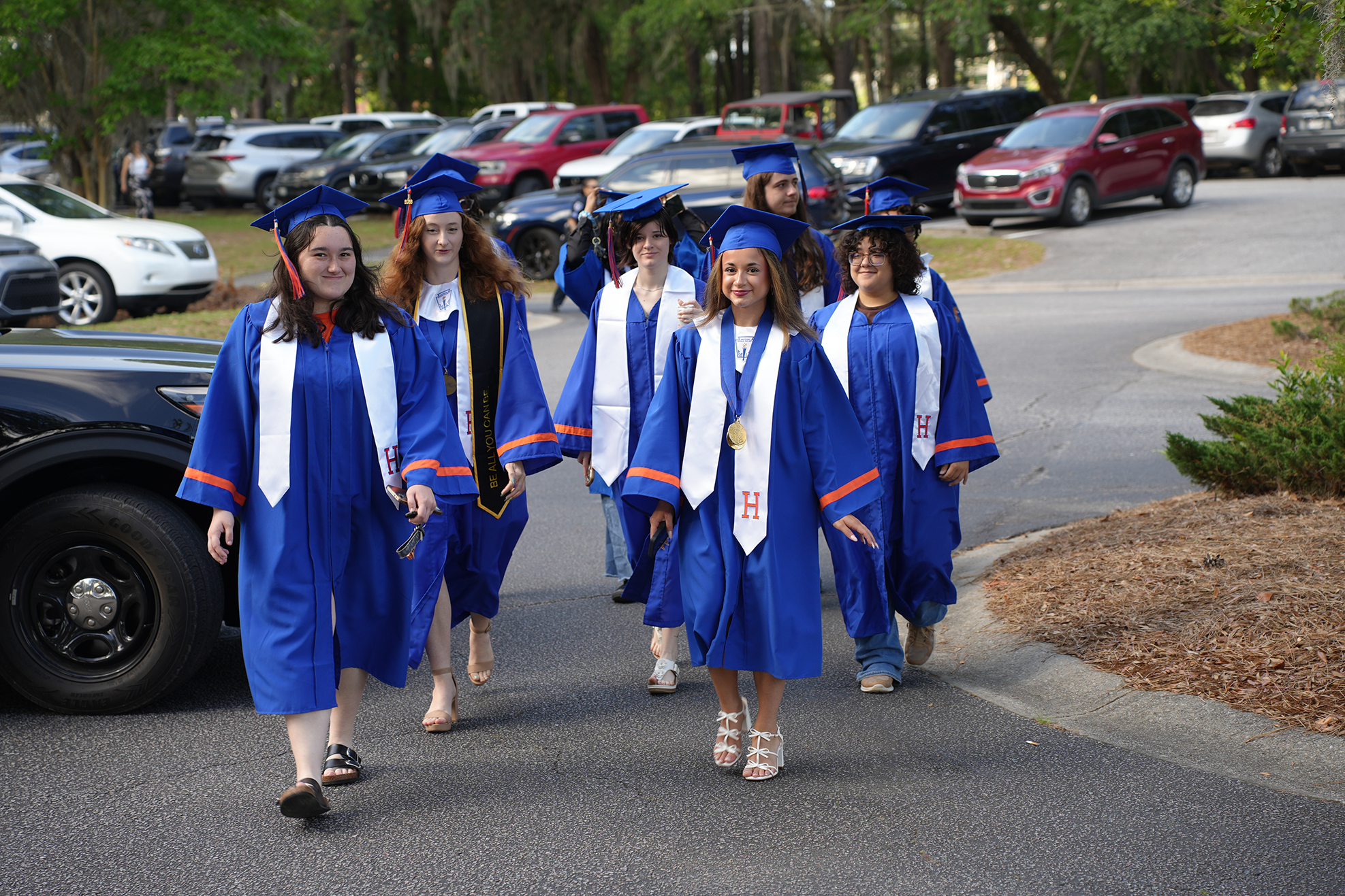 A group of six people in blue graduation gowns walking in a parking lot. Several cars parked in the background.