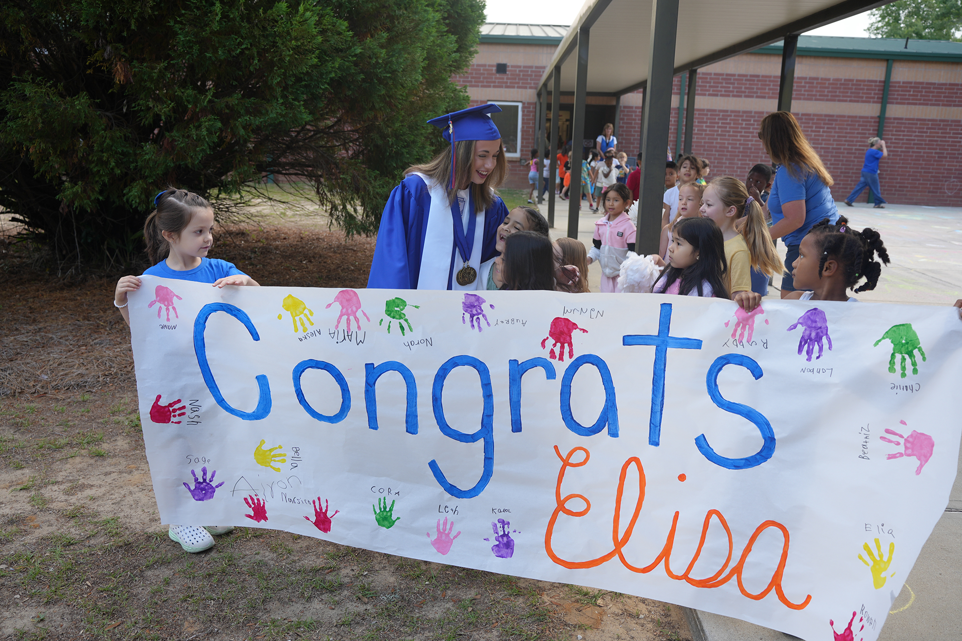 Graduates in caps and gowns hold a banner with colorful handprints. It reads "Congrats Elisa." People gather in a school setting.
