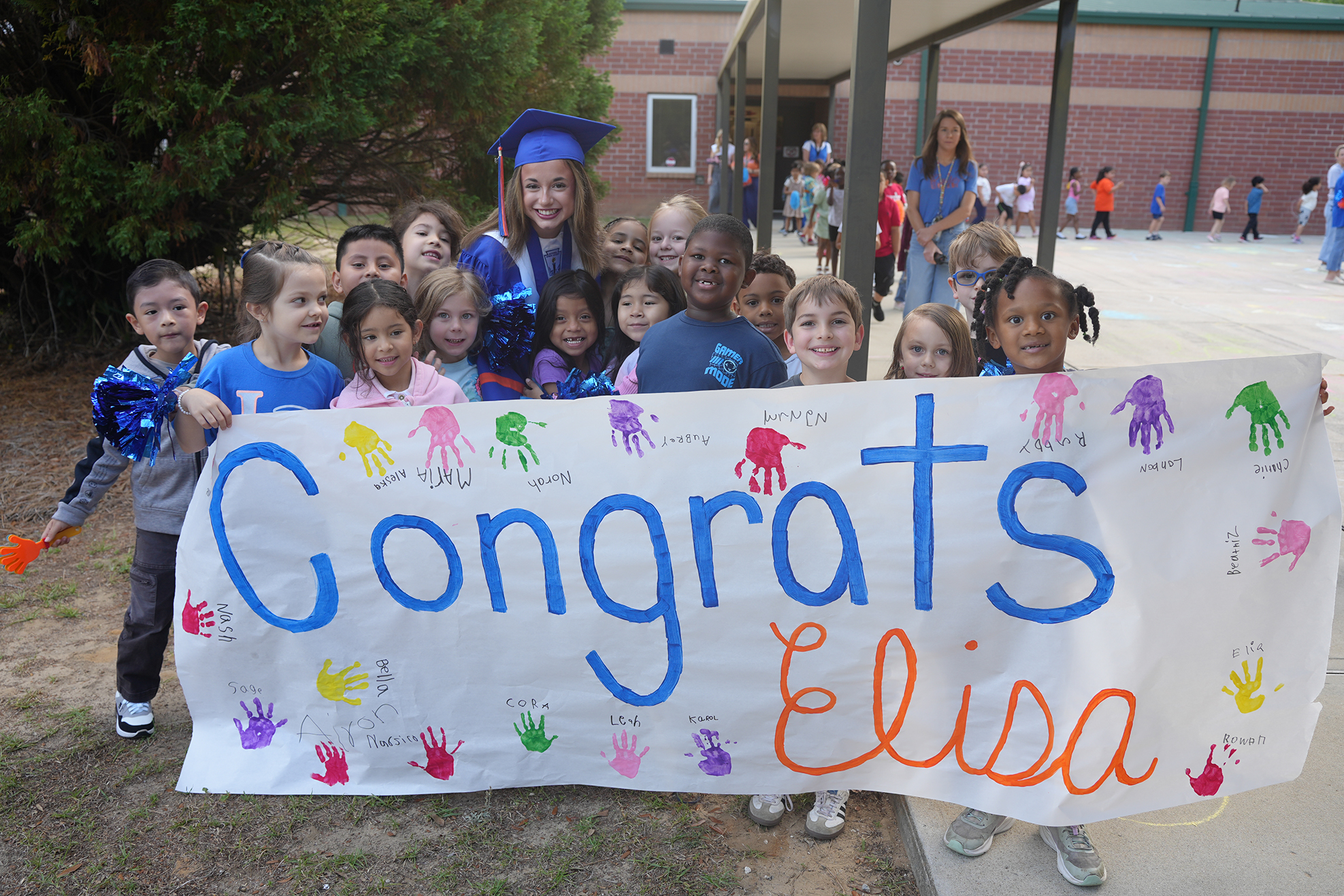 A group of children holding a banner that says "Congratulations, Elisa" stand outside a school building.