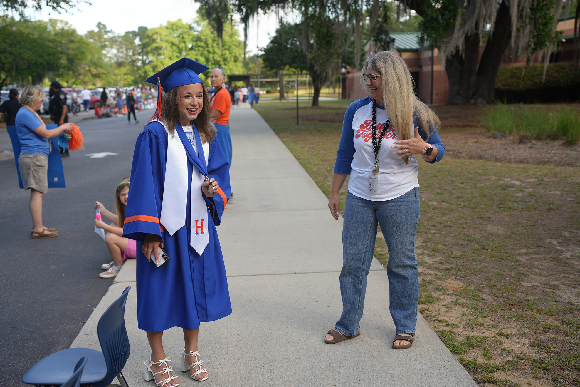 A woman in a blue gown stands next to a woman in a white shirt. Behind them, a sidewalk leads to parked cars.