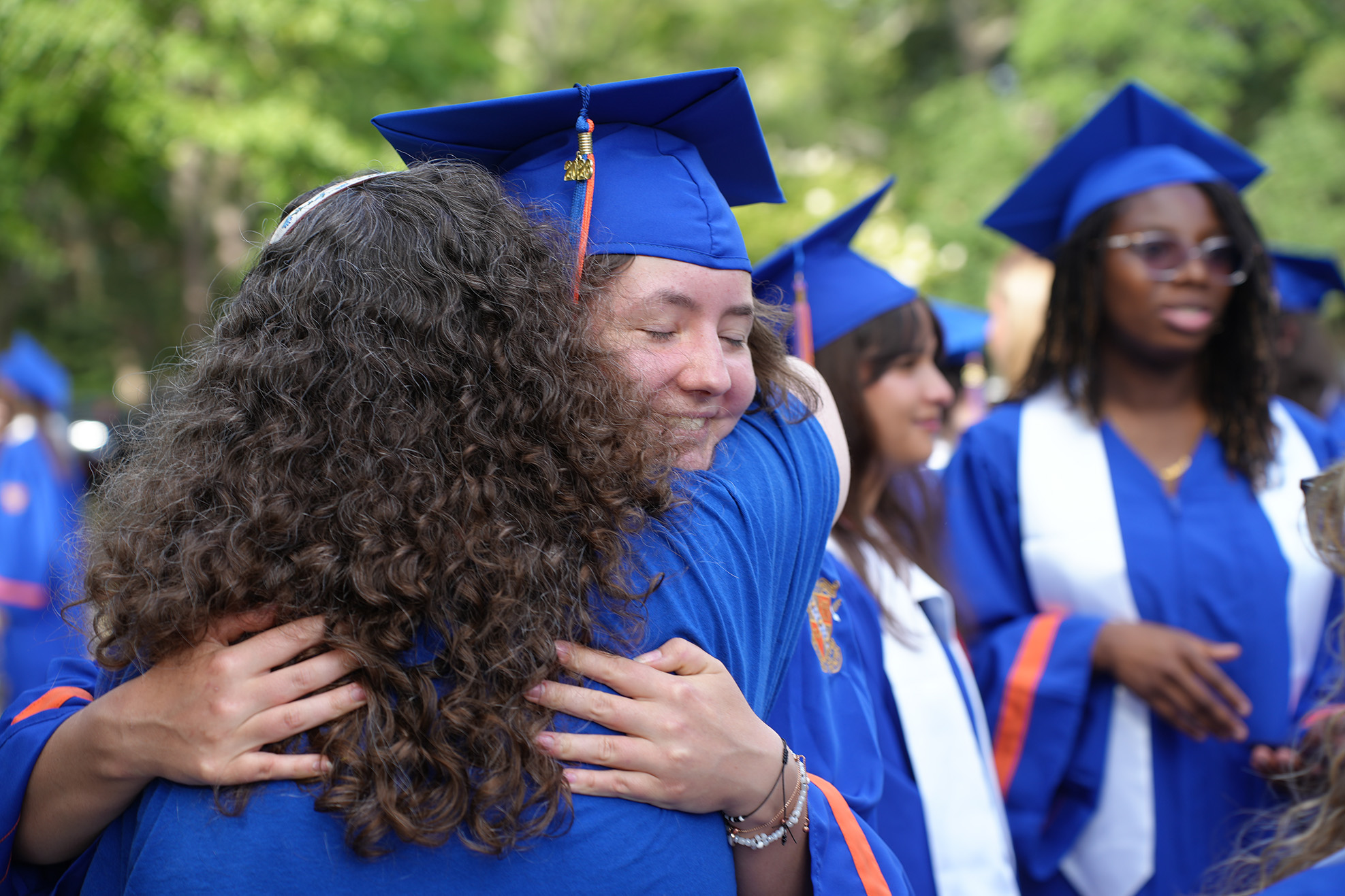 Group of graduates in blue gowns and caps, one hugging a woman with curly hair. Blurry trees and other students in the background.
