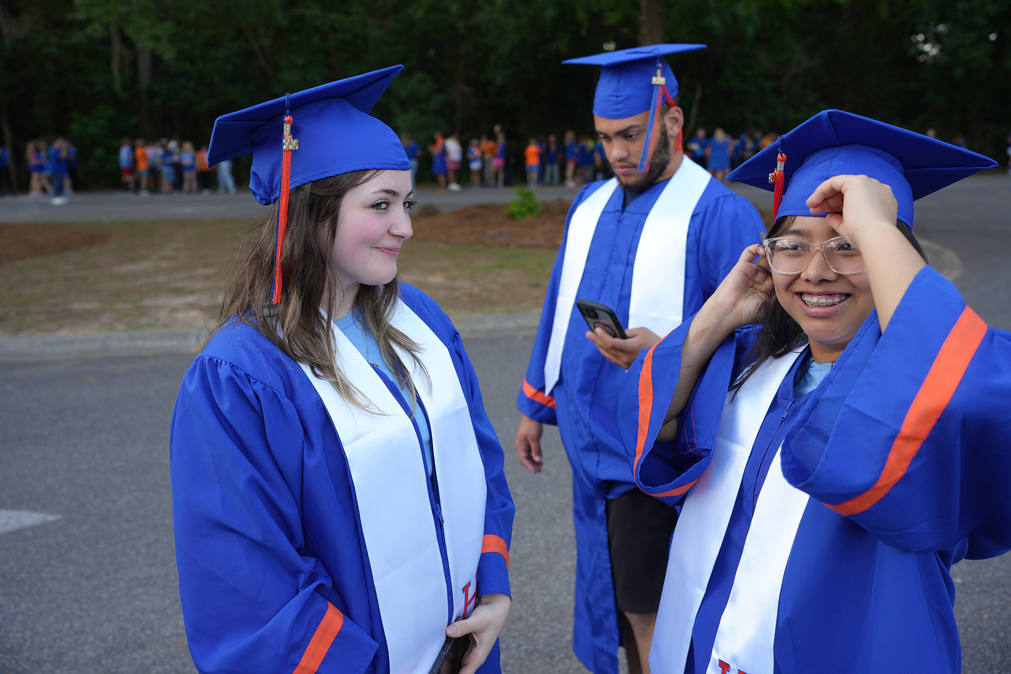 Three graduates in blue gowns and caps stand in a parking lot, one adjusting her hat.