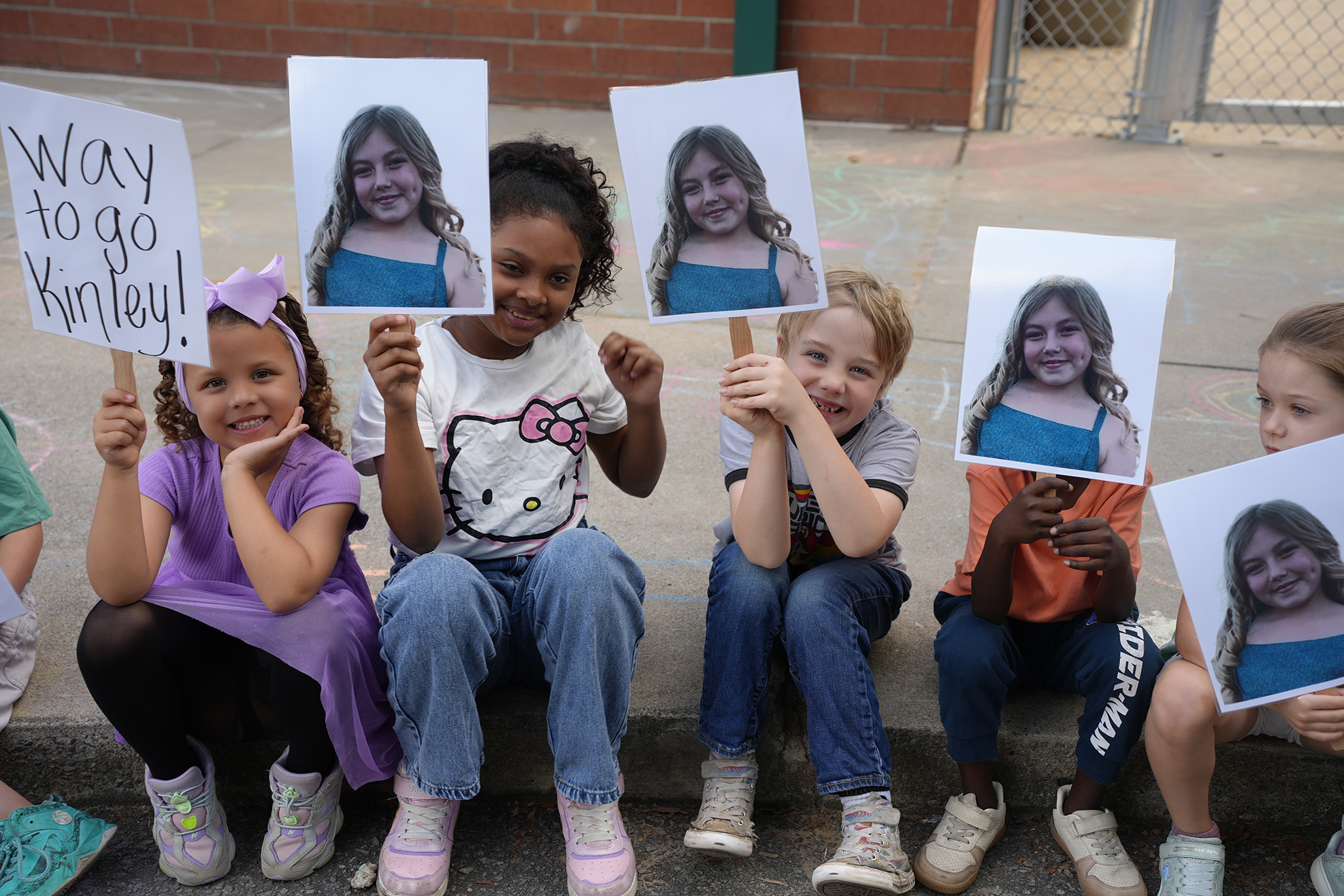 Group of children sitting on a curb, each holding signs with a picture of a girl.