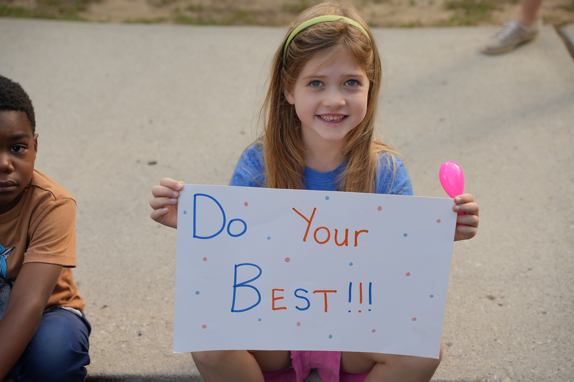 A young girl holding a sign that says "Do Your Best!!!" with a balloon, seated next to another child.