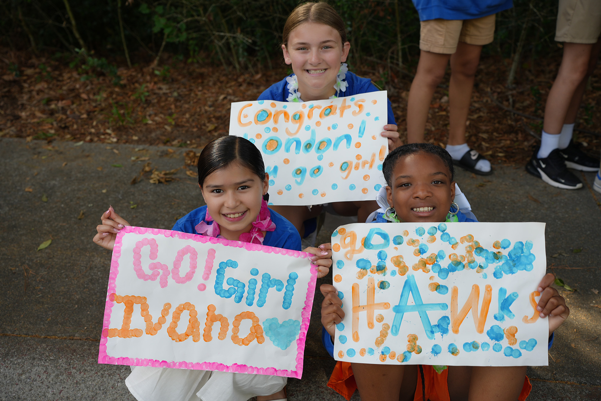 Three girls sit on the ground, holding handmade signs. The front girl's sign reads "Go! Girl Ivana". Background shows greenery and people.