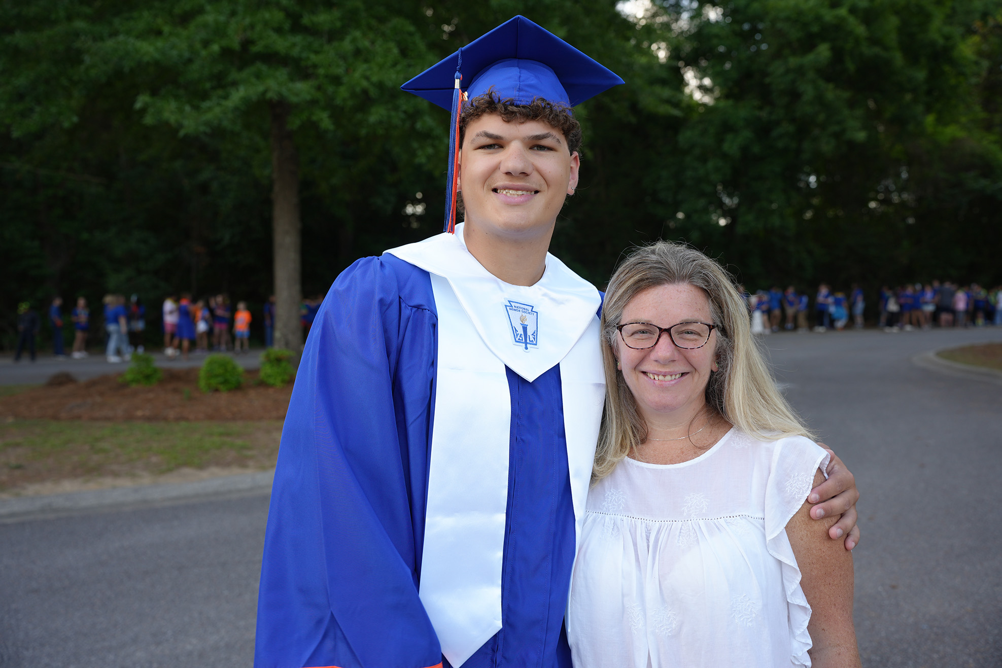 A person in a blue gown and cap stands next to a person in white. Behind them, trees and a blurred crowd in the distance.