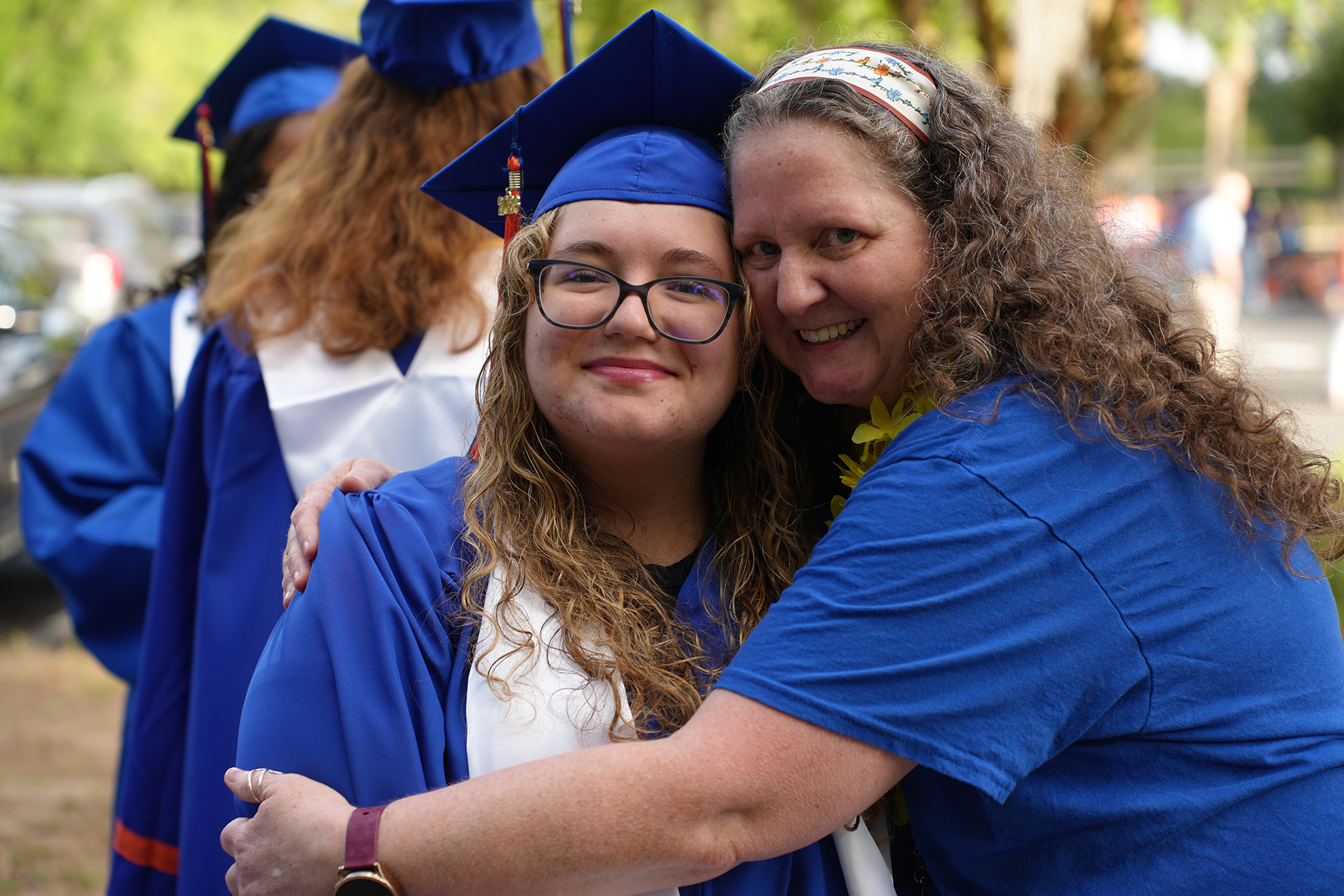 A woman in a blue cap and gown hugs another person, both wearing blue shirts. Behind them, a few people in blue graduation gowns.