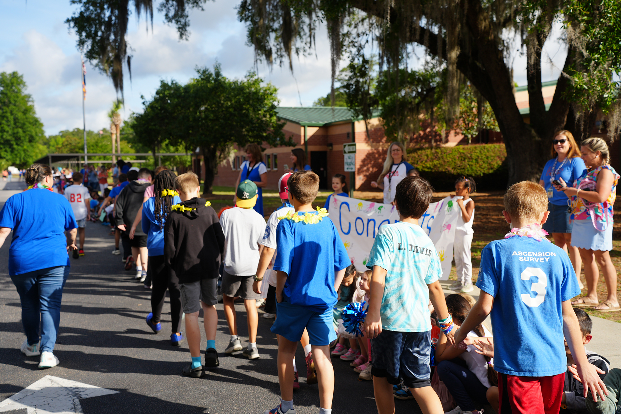 A group of people in blue and red shirts walk down a street with greenery. Some hold a white banner.