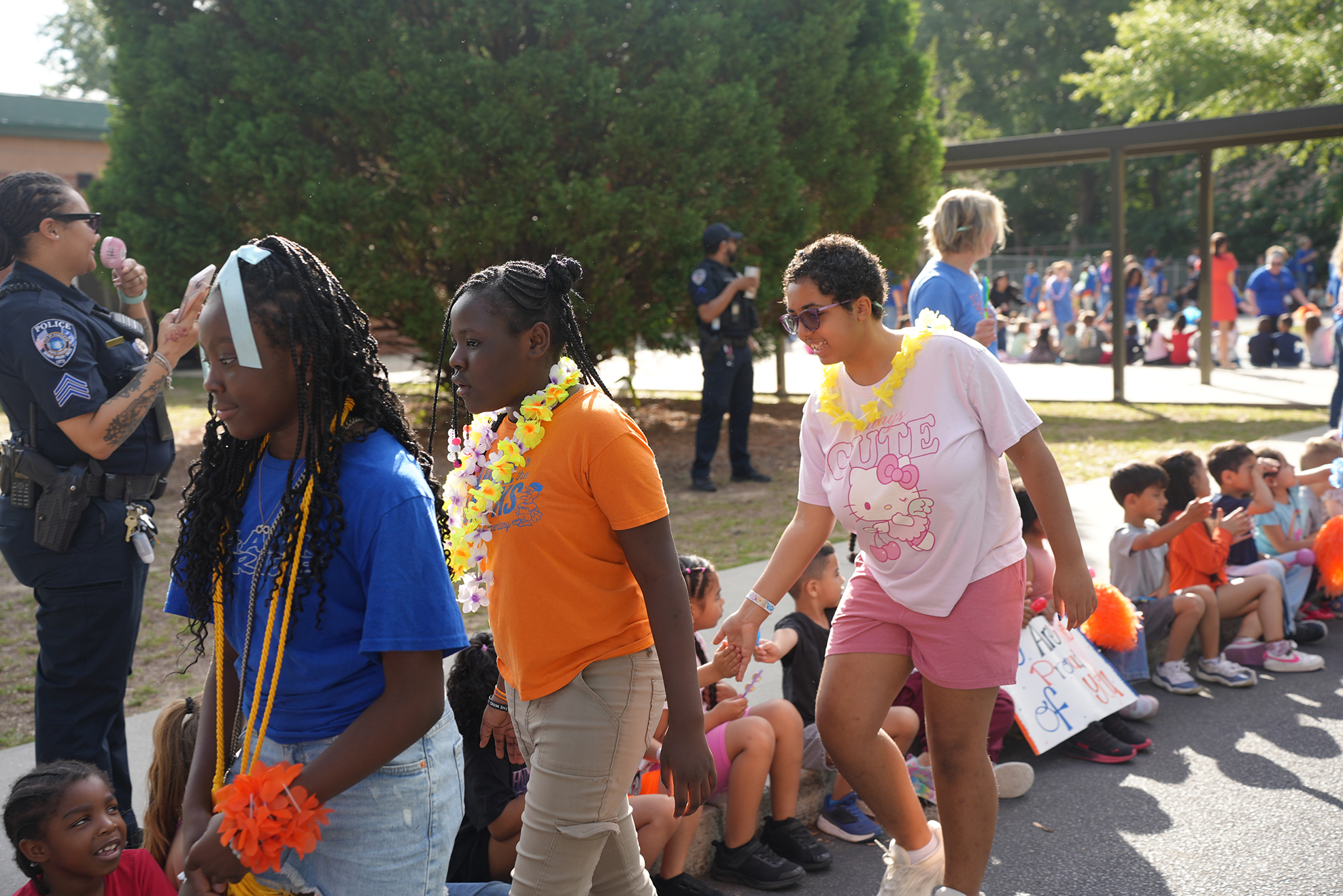 Group of children in schoolyard with two adults, one wearing police uniform, all wearing festive accessories.