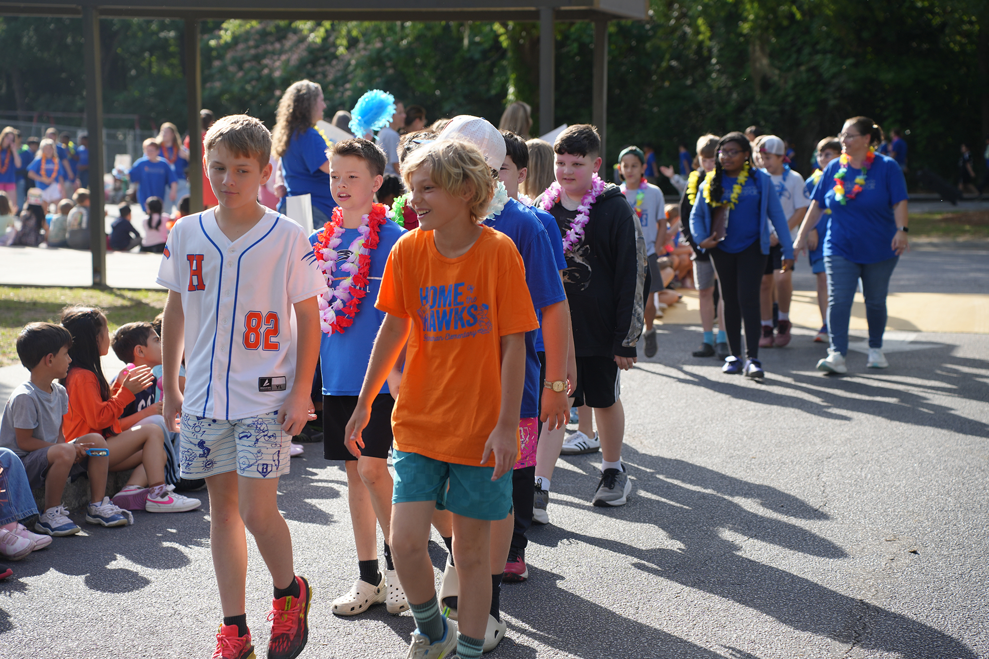 Kids in casual attire, some wearing leis, walk in an outdoor area. Some sit on the ground.