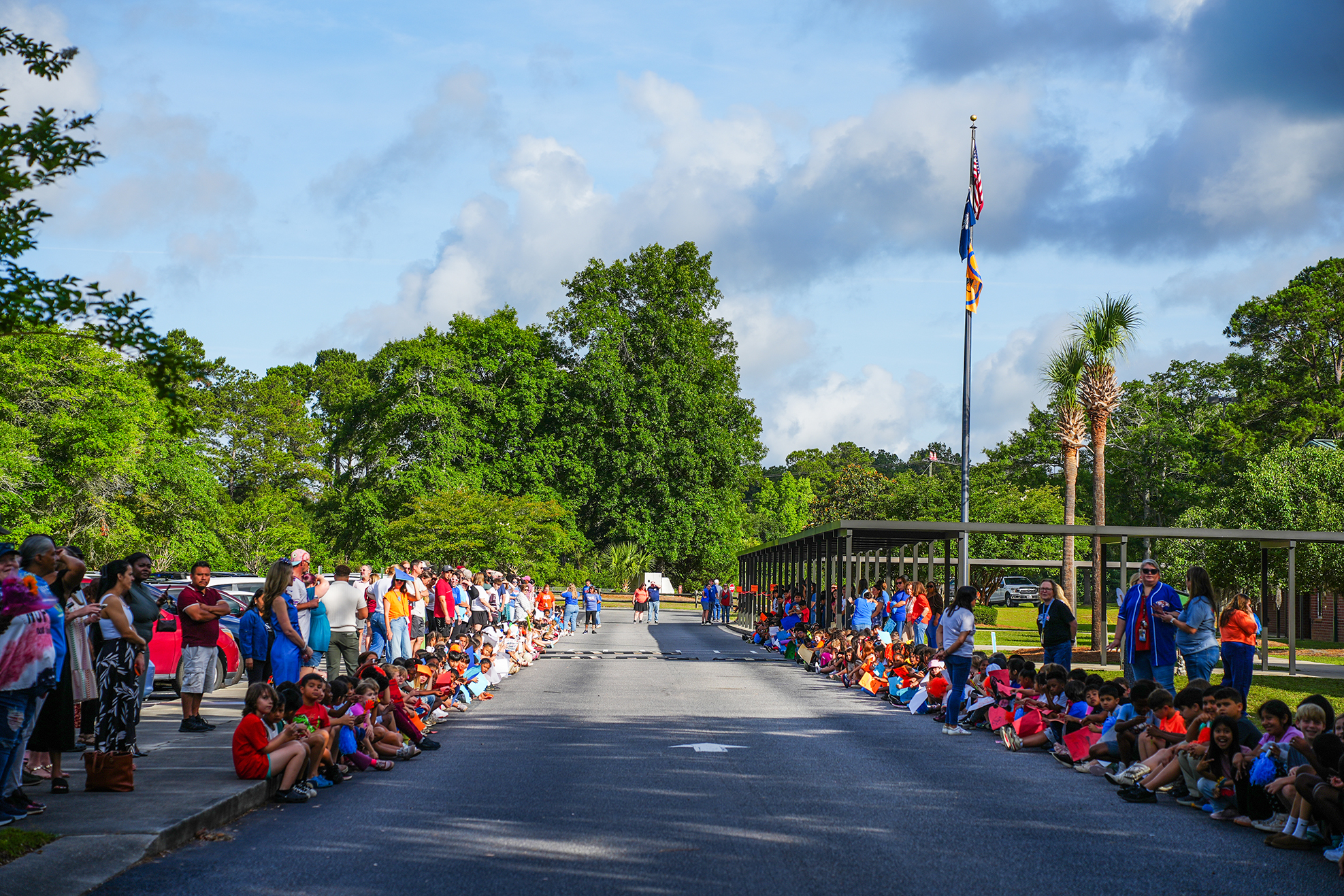 People line the road, with some standing and others sitting. A flag is on a pole near a canopy. Trees are in the background.