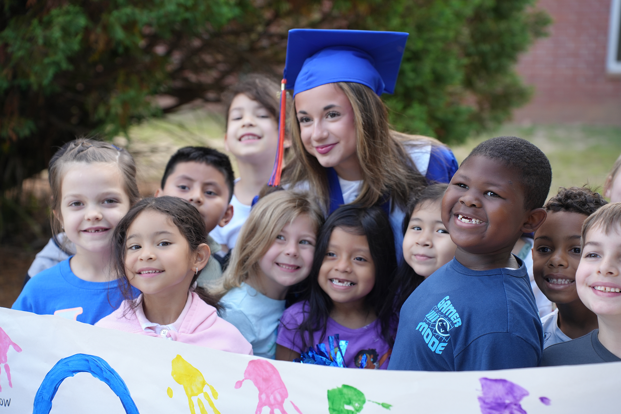 Group of children in front of a colorful banner, including a graduate, standing with a building in the background.