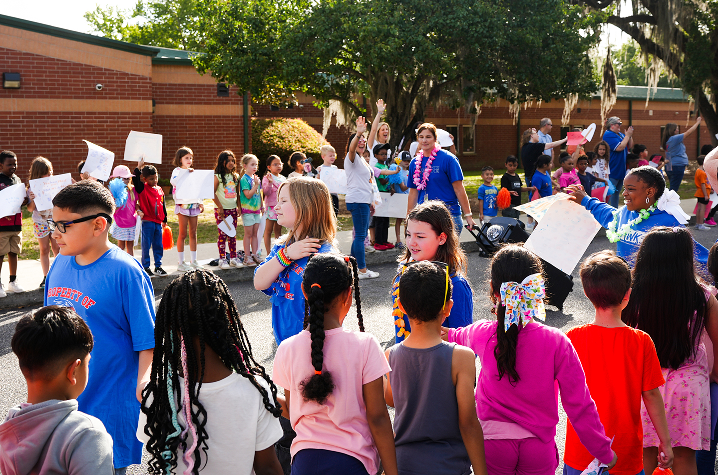 Children in blue shirts gather in front of a school building, holding signs and engaging with adults.