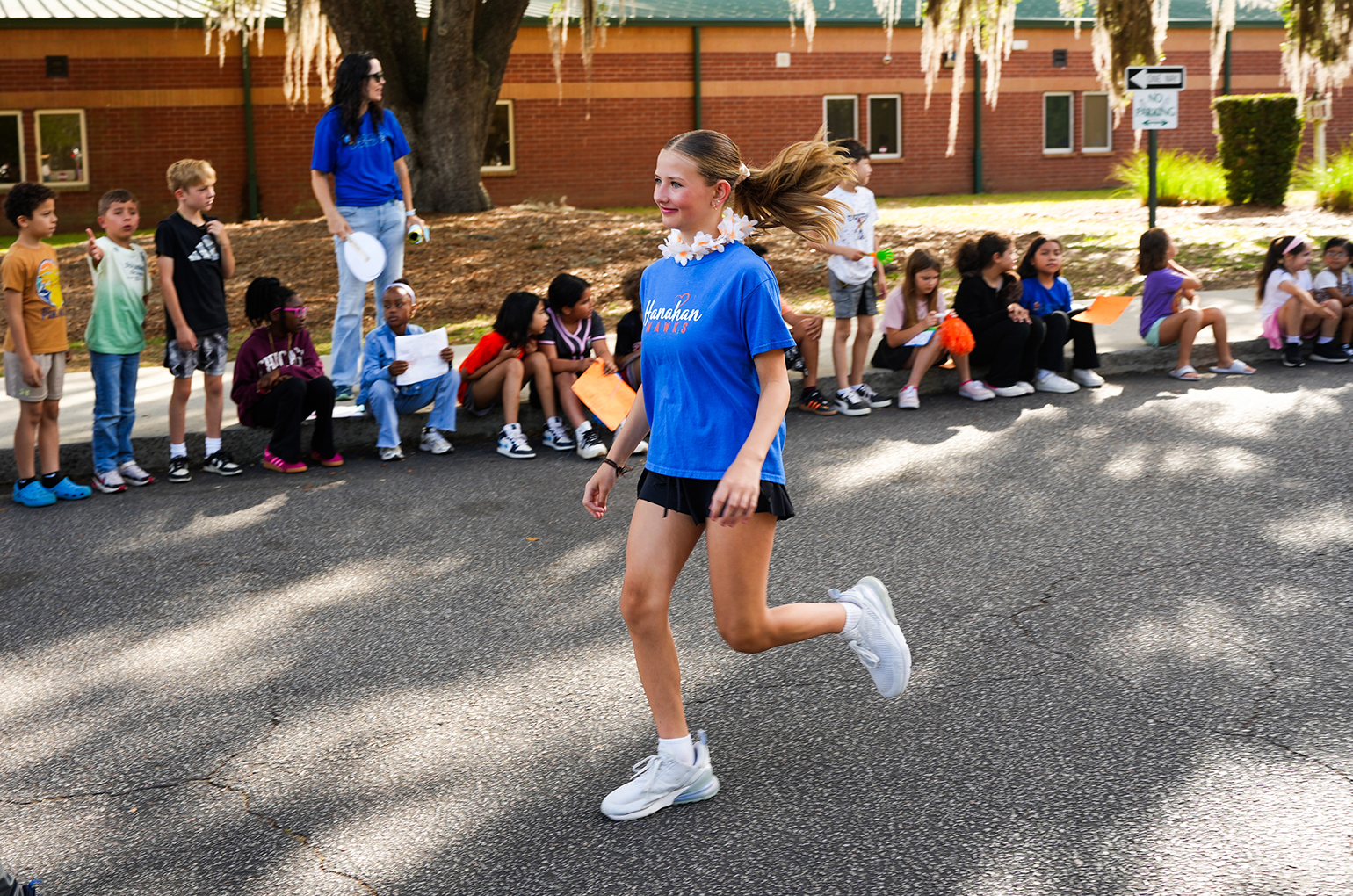 A young girl runs on a street while children sit on a curb, some holding paper. A building with brick walls is in the background.