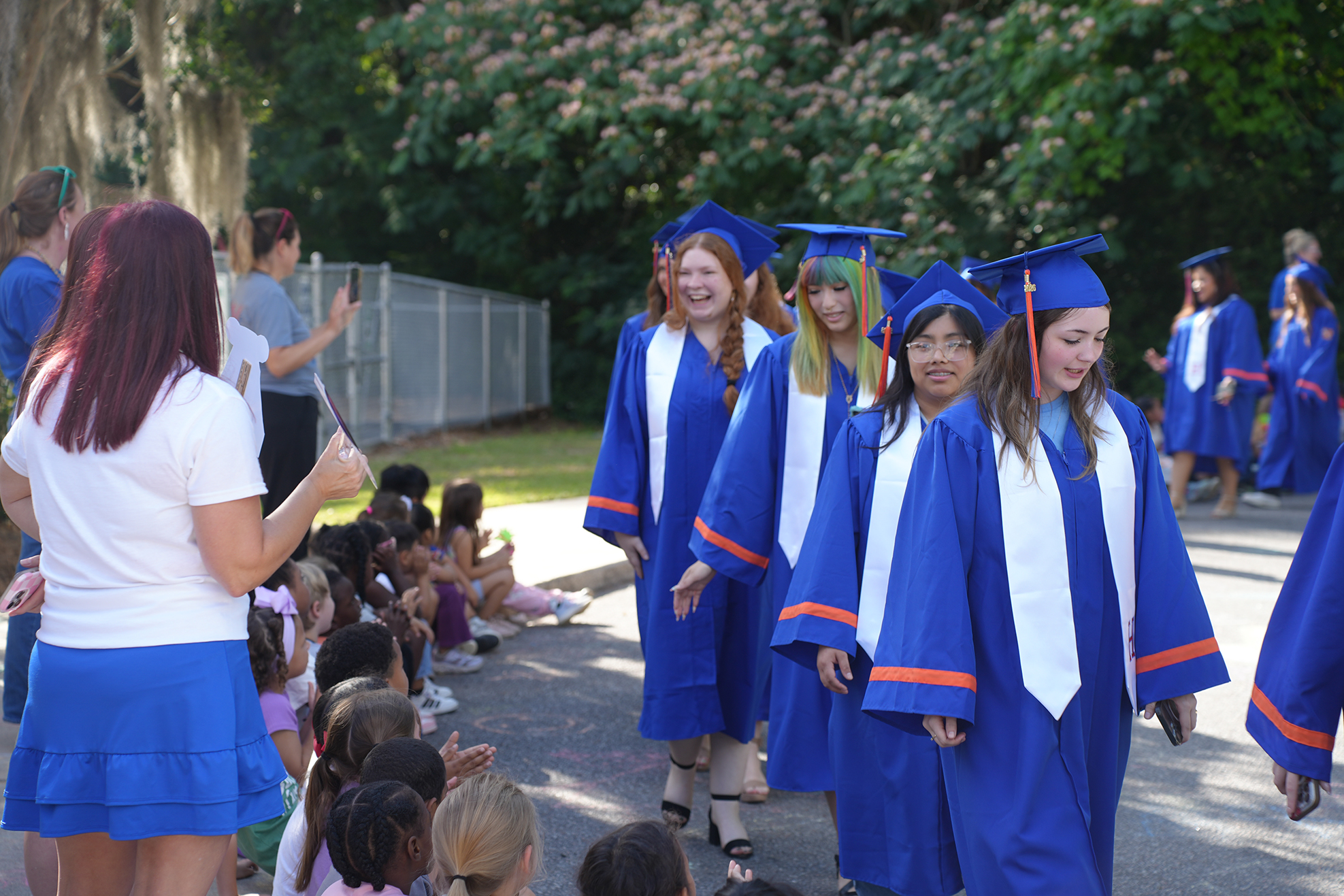 Graduates in blue gowns with white sashes and blue hats walk through a crowd. Some wear red hair.