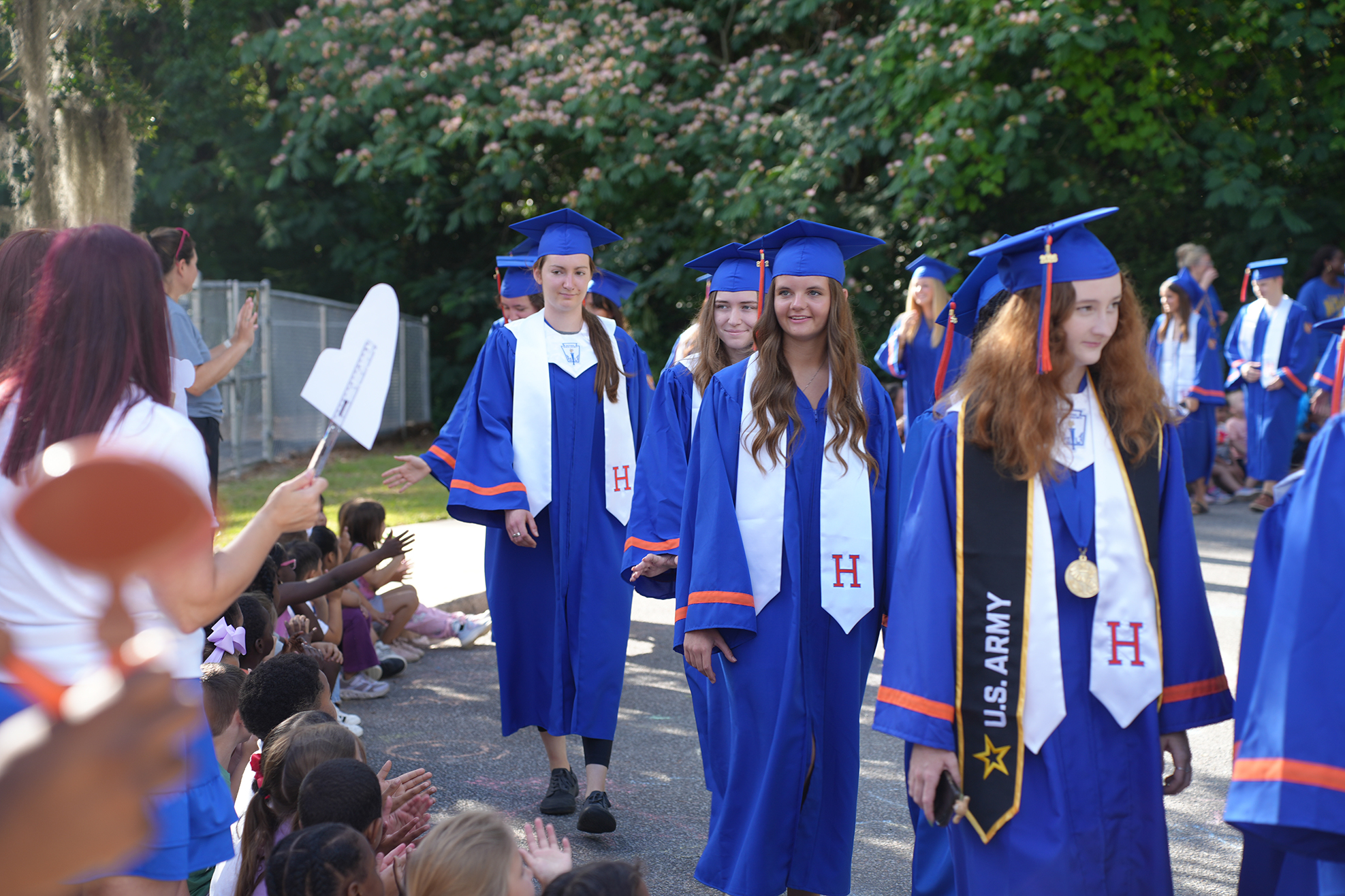 Graduates in blue gowns walk past a crowd of children, one wearing a medal and sash.
