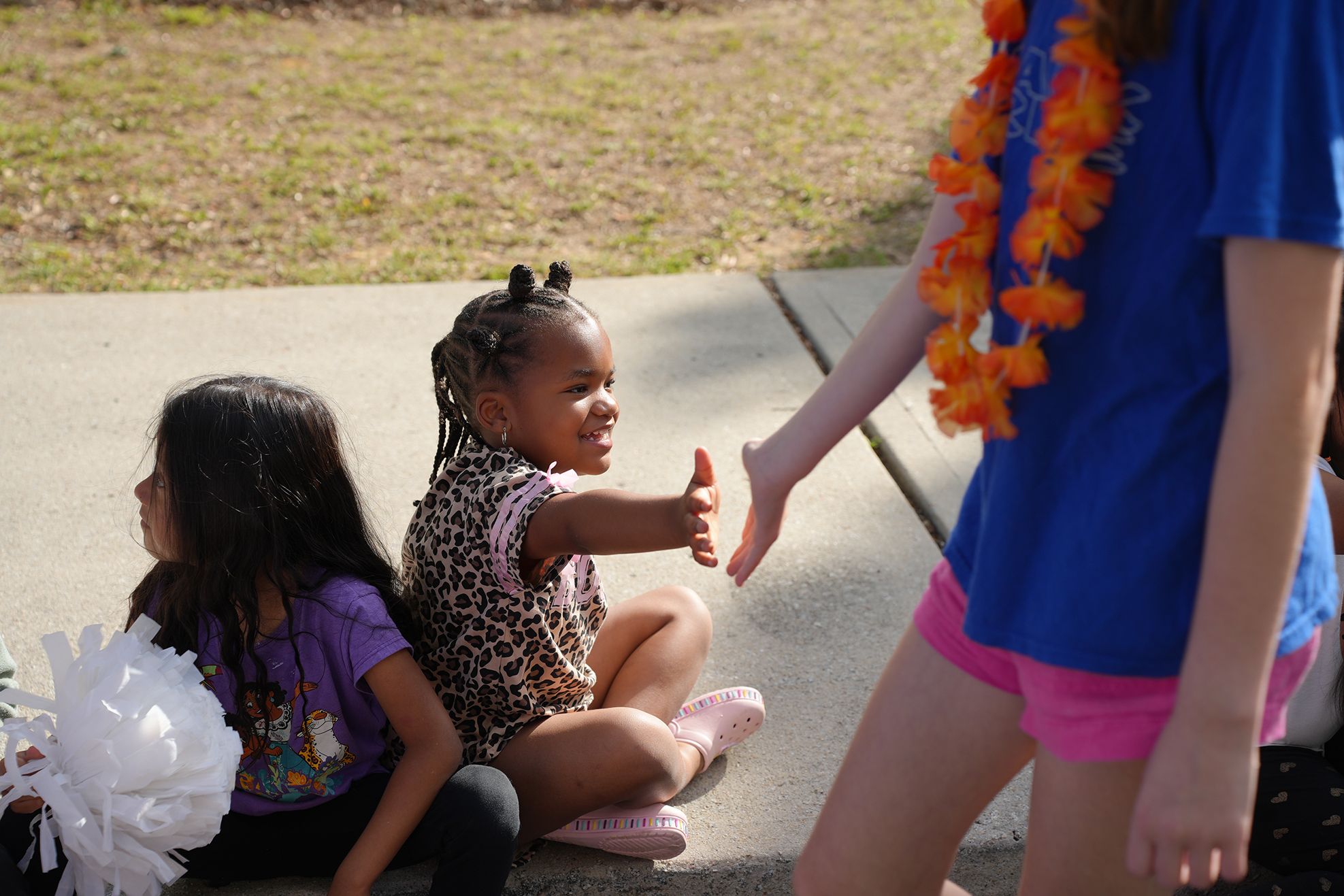 Two children sit on the ground, one wearing a floral shirt. Another child approaches, wearing a blue shirt, pink shorts, and a lei.