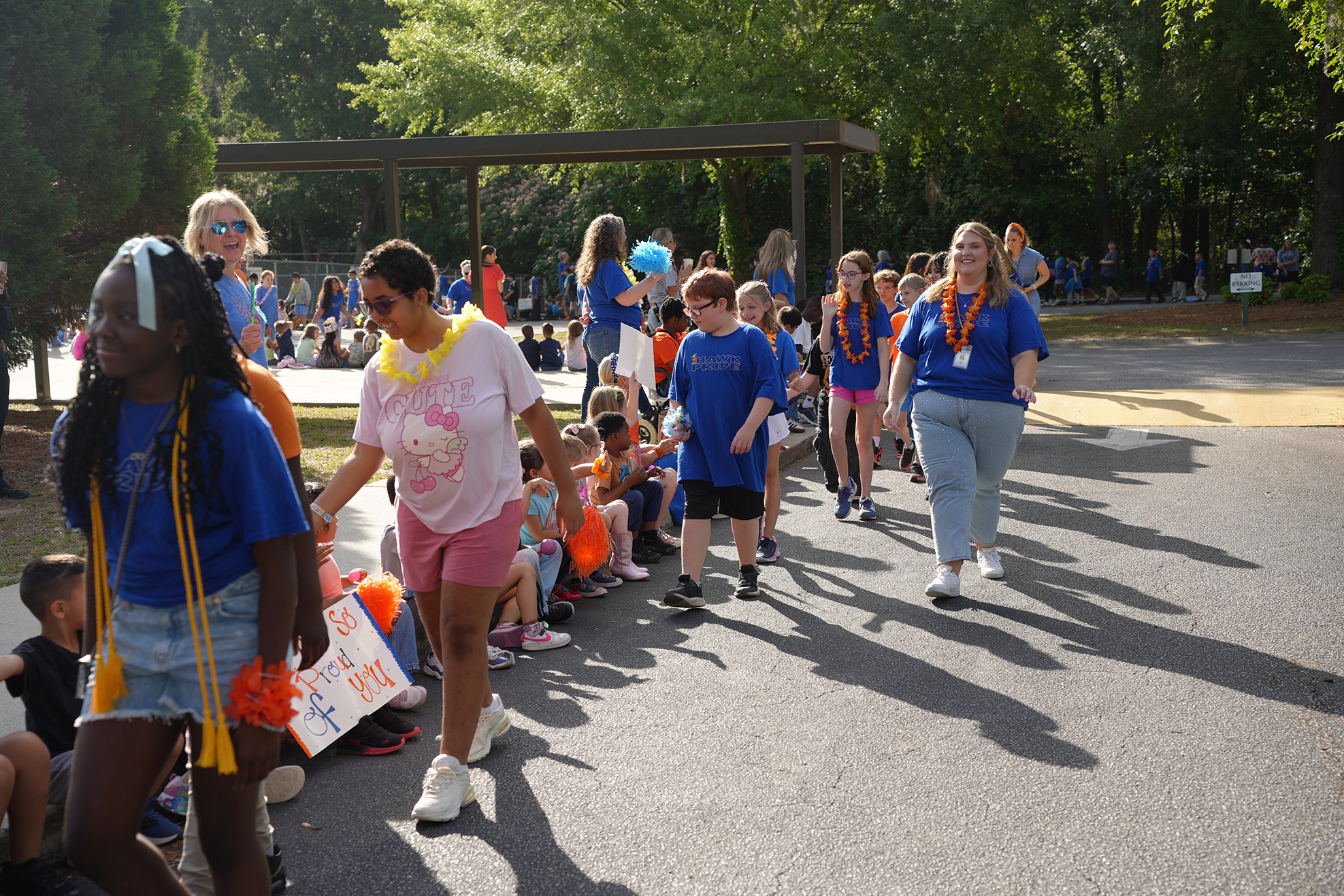 A group of people in blue shirts walk on a paved road with trees in the background.